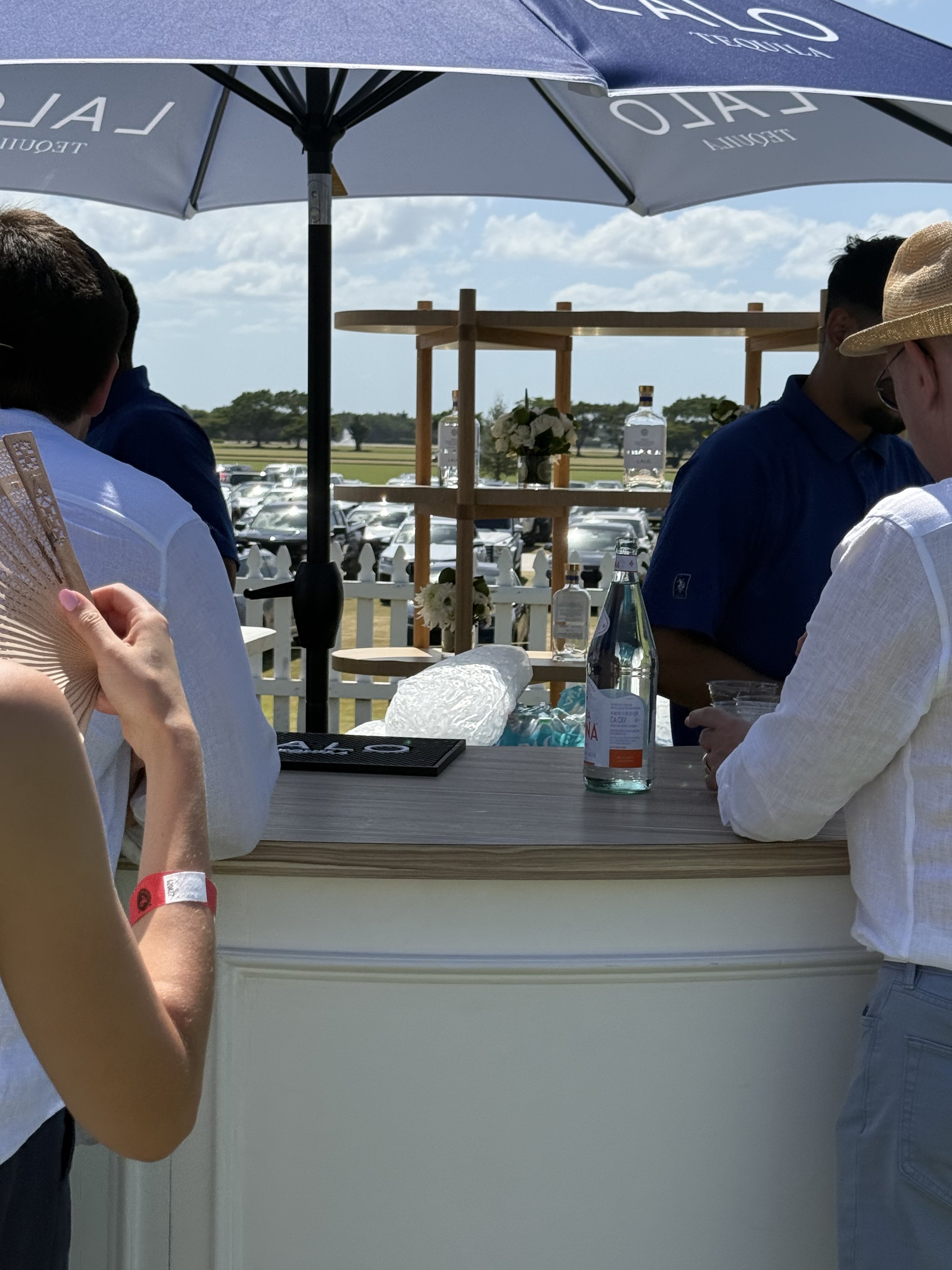 People gathered at a beverage stand under a large umbrella, with a scenic outdoor setting, including a parking lot, a field, and trees, in the background.