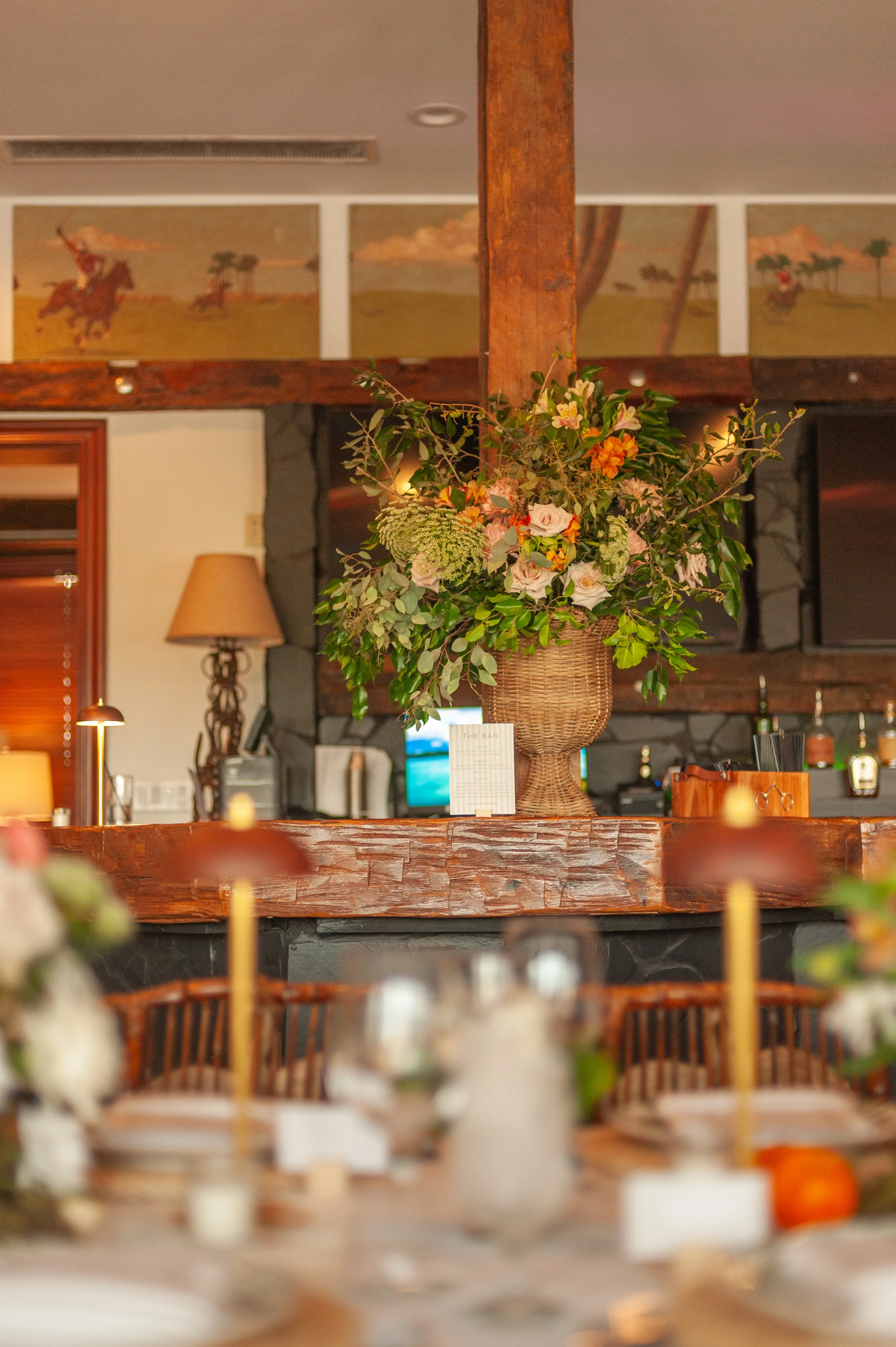 A large floral arrangement in a wicker basket on a wooden bar counter in a cozy restaurant or cafe. The background features stone and wood decor with paintings and warm lighting.