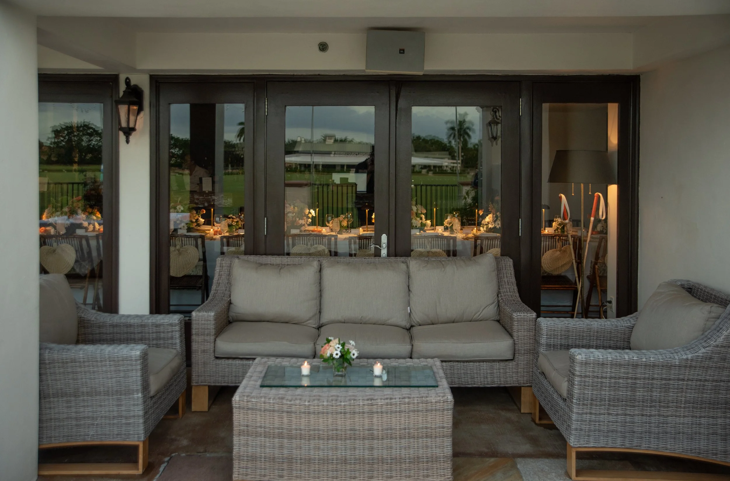 Indoor patio area with beige wicker furniture, including a sofa and two armchairs, with candles and a flower centerpiece on a glass-topped coffee table. Behind the furniture, there are glass double doors leading to an outdoor dining area with tables 