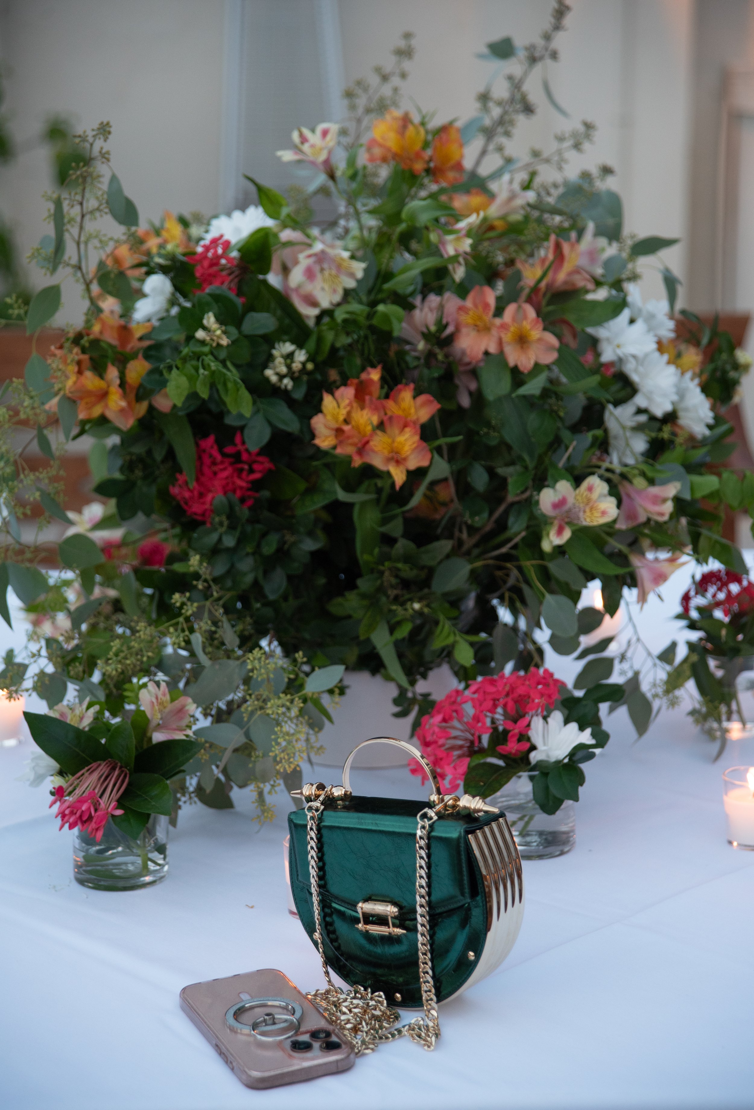 A large floral arrangement in a white vase with pink, orange, white, and green flowers and greenery, placed on a white table. In front of the flowers, there is a small green handbag with gold accents, a gold chain strap, a pink phone with a ring hold
