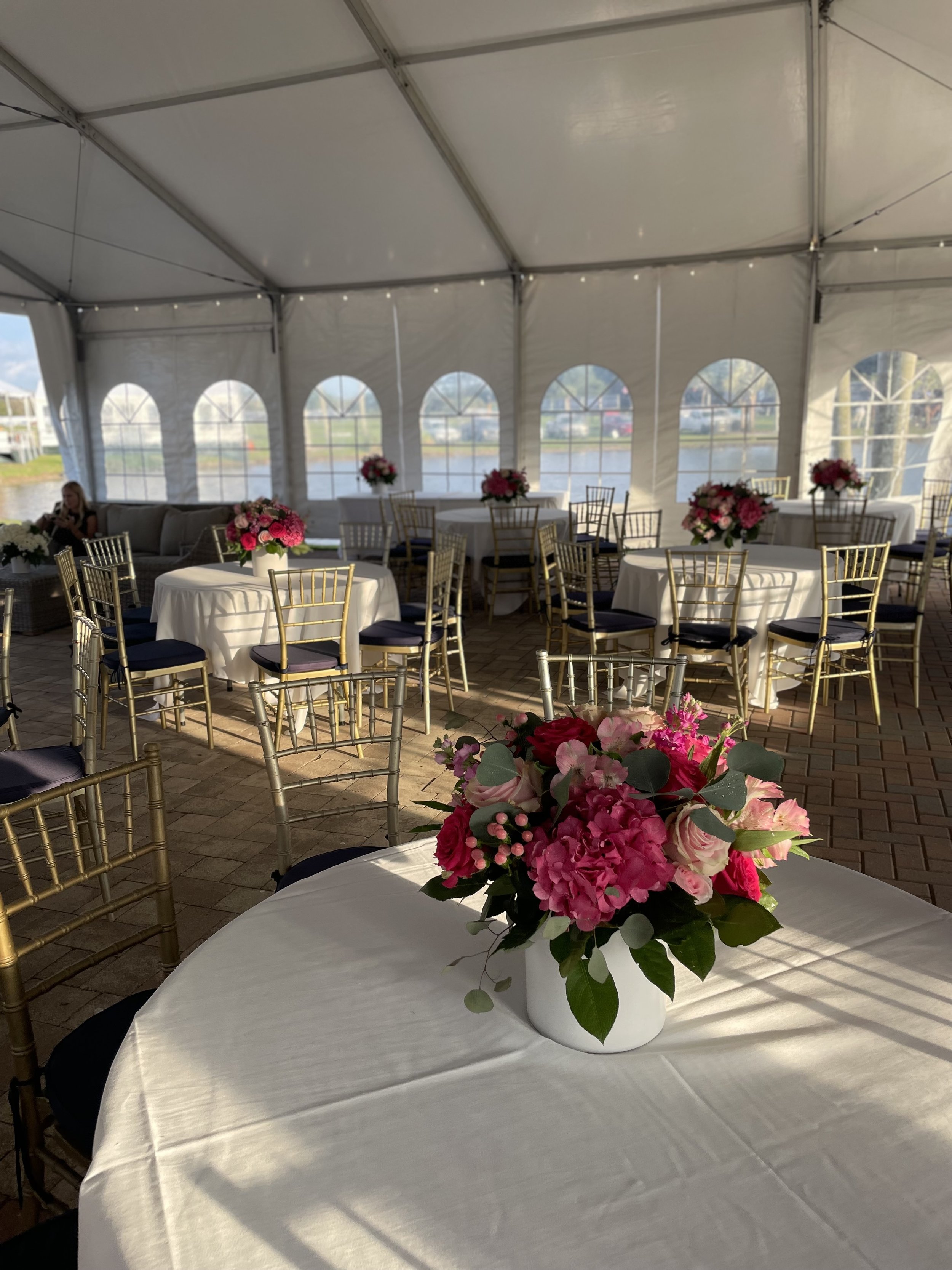 Inside a decorated event tent with round tables adorned with pink floral centerpieces, gold chairs, and a brick floor.