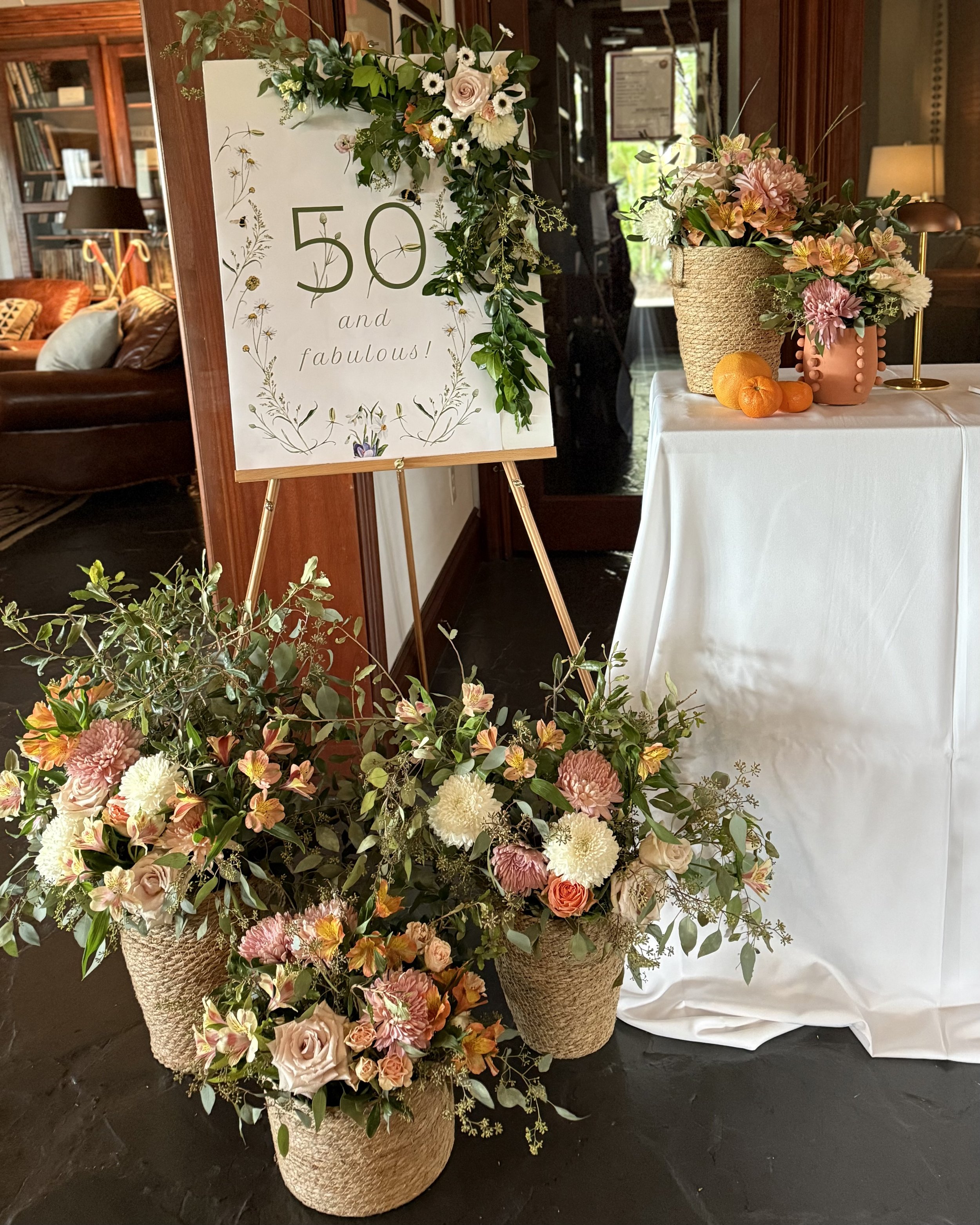 Decorative floral arrangement with roses, dahlias, and greenery in woven baskets, a white tablecloth, a sign on a wooden easel celebrating a 50th birthday with the words '50 and fabulous!', and a small orange fruit on the table.