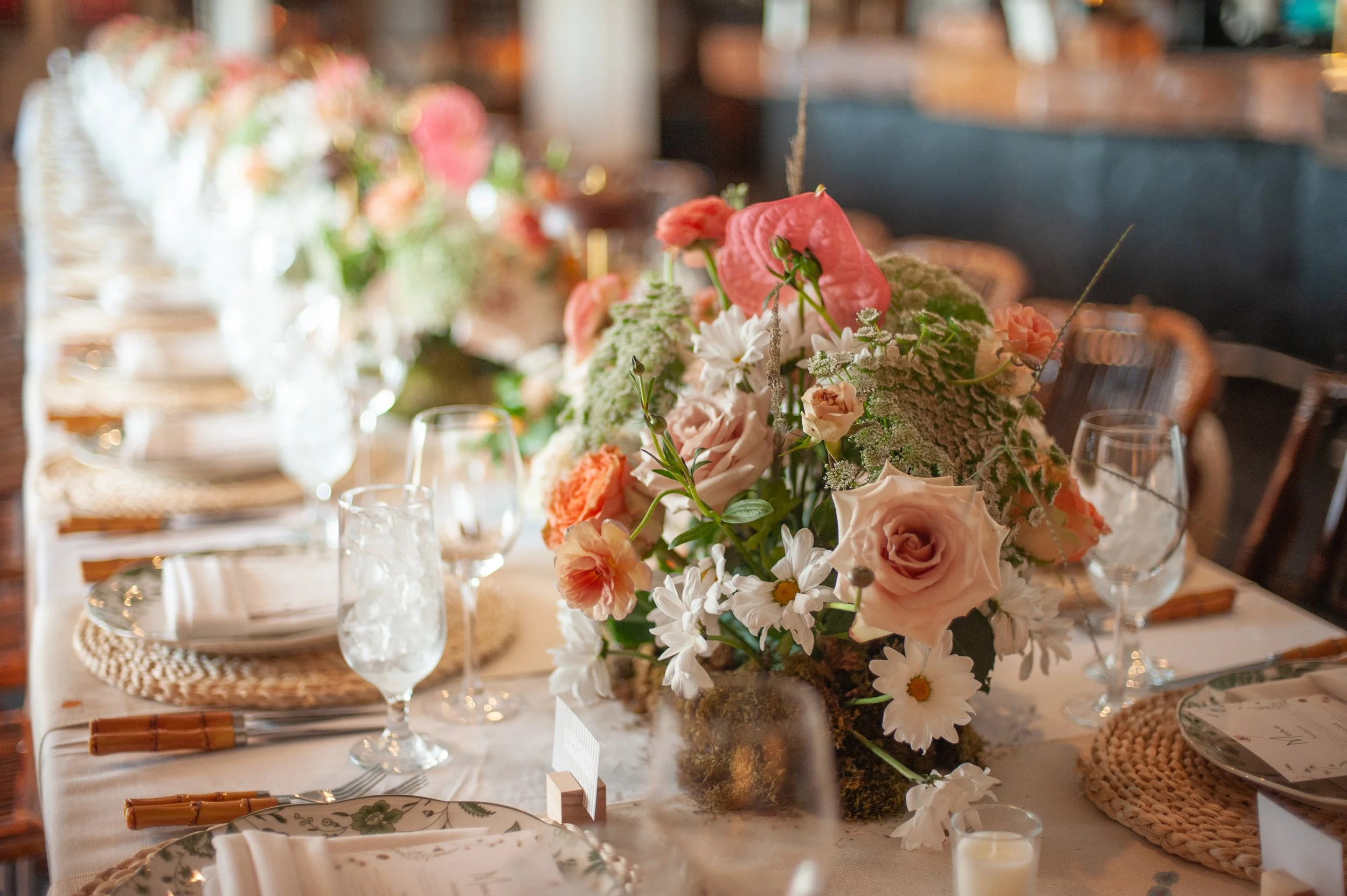 A long dining table decorated with a large floral centerpiece containing roses, daisies, and other flowers. The table is set with plates, glasses, and napkins in a restaurant or event space.