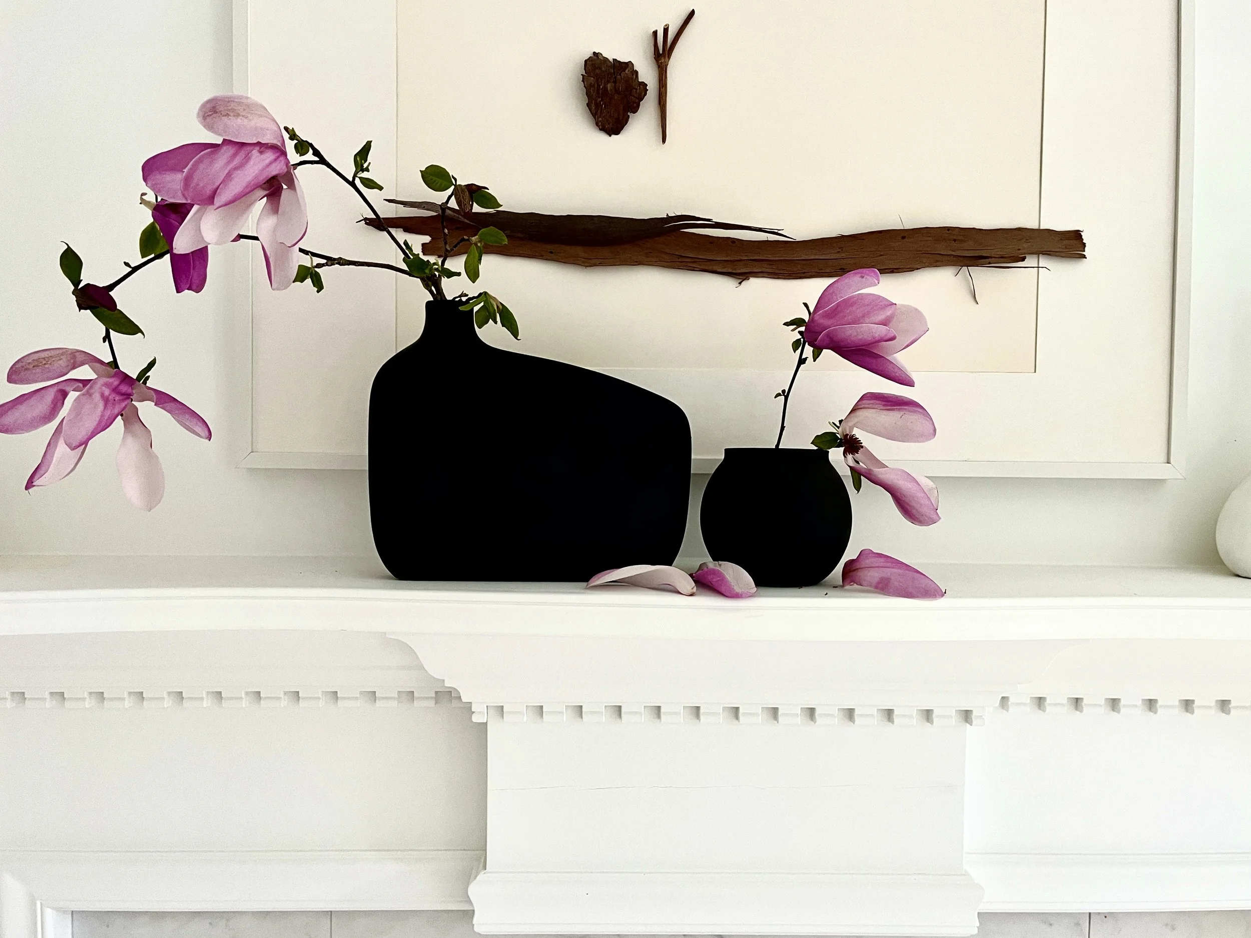 Two black vases with pink magnolia flowers and petals, arranged on a white fireplace mantel with decorative molding, with a wall art piece made of driftwood and sticks in the background.