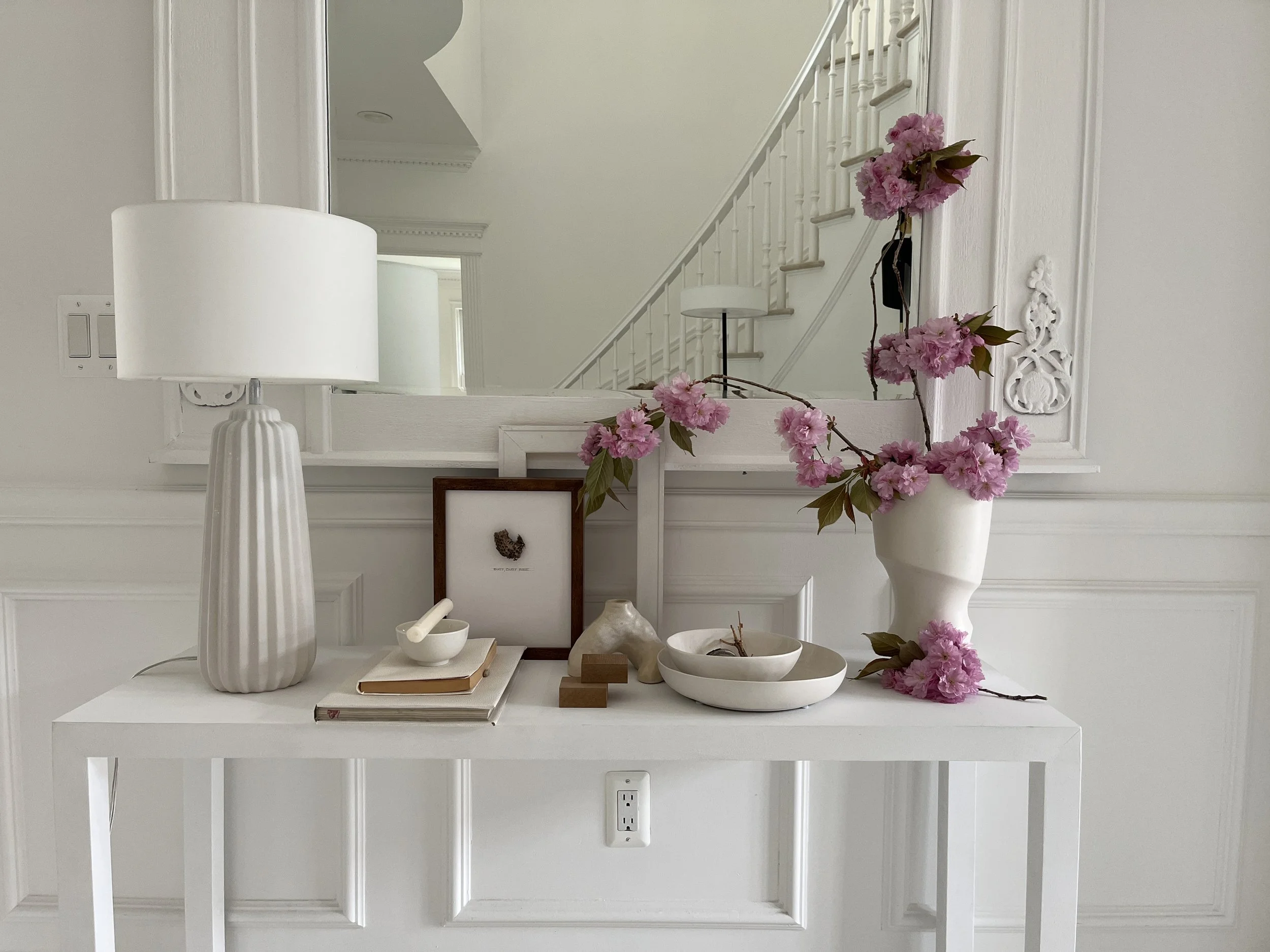 A white console table with a large lamp, framed artwork, bowls, a ceramic dog sculpture, and a vase with pink flowers in a white room with molding and a mirror.