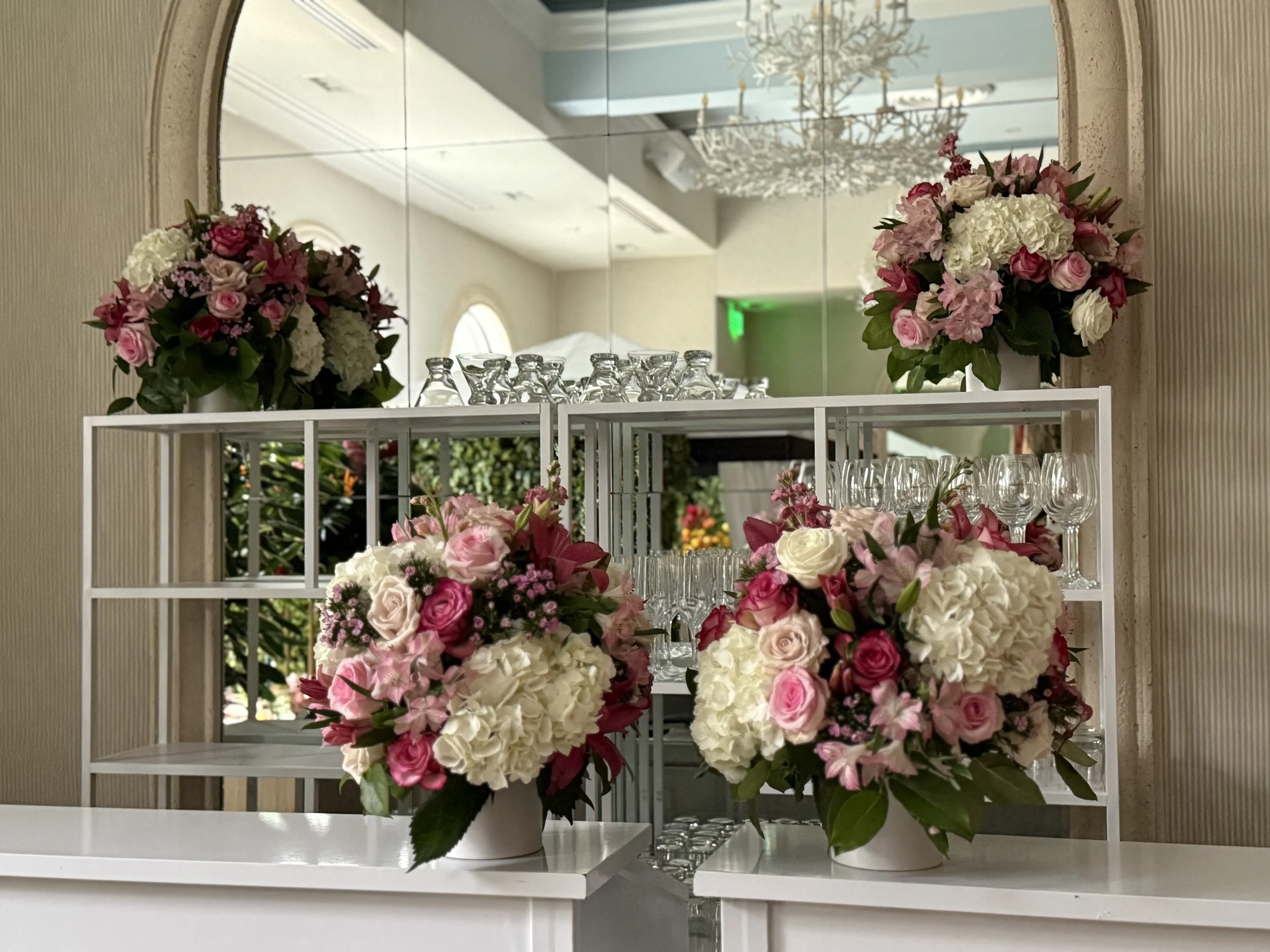 Elegant floral arrangements with pink and white roses, white hydrangeas, and greenery on a white table, with a mirror, wine glasses, and a chandelier reflected in the background.