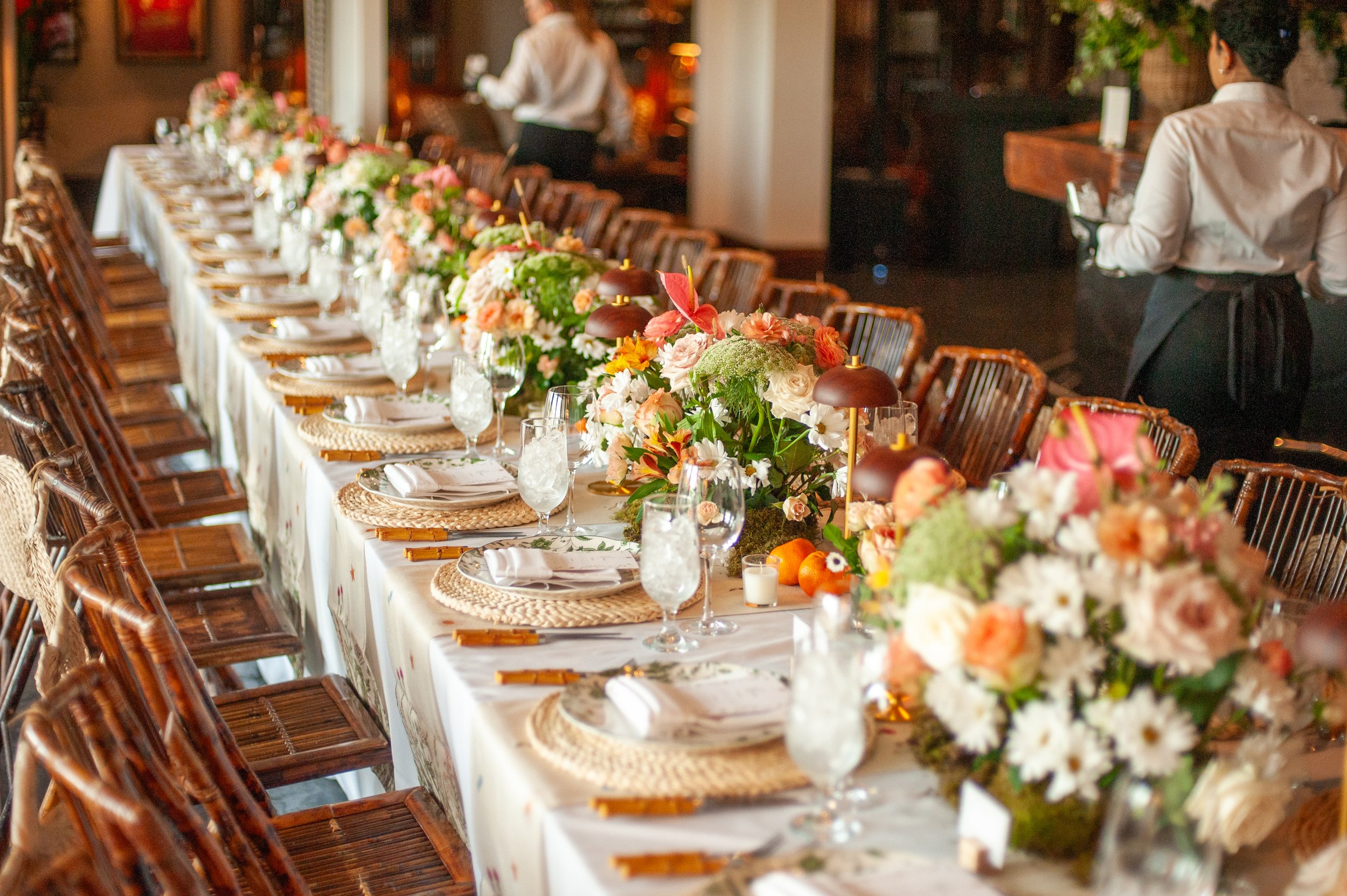 Long wedding or banquet table decorated with floral arrangements, candles, and dinnerware, set in a warmly lit indoor space with waitstaff attending.