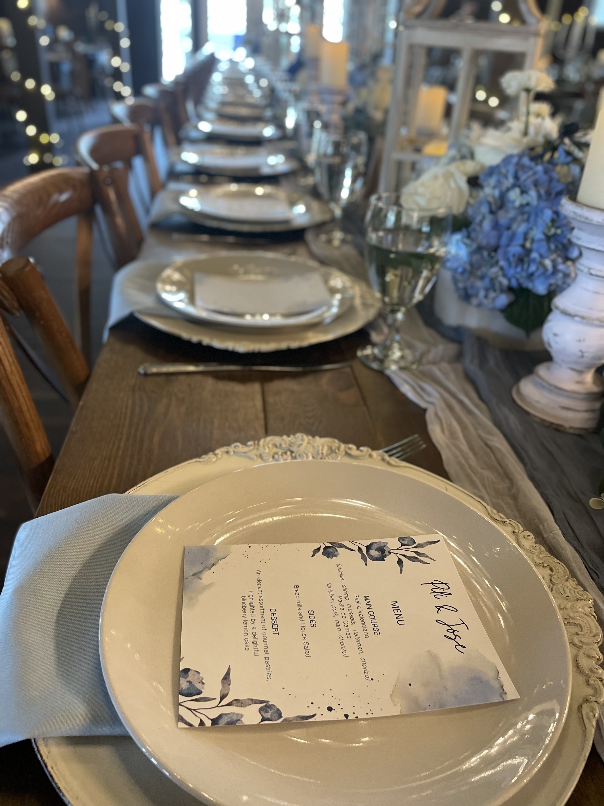Elegant dinner table set with white plates, silverware, wine glass, and menu card on a ceramic plate, with floral centerpiece and string lights in the background.