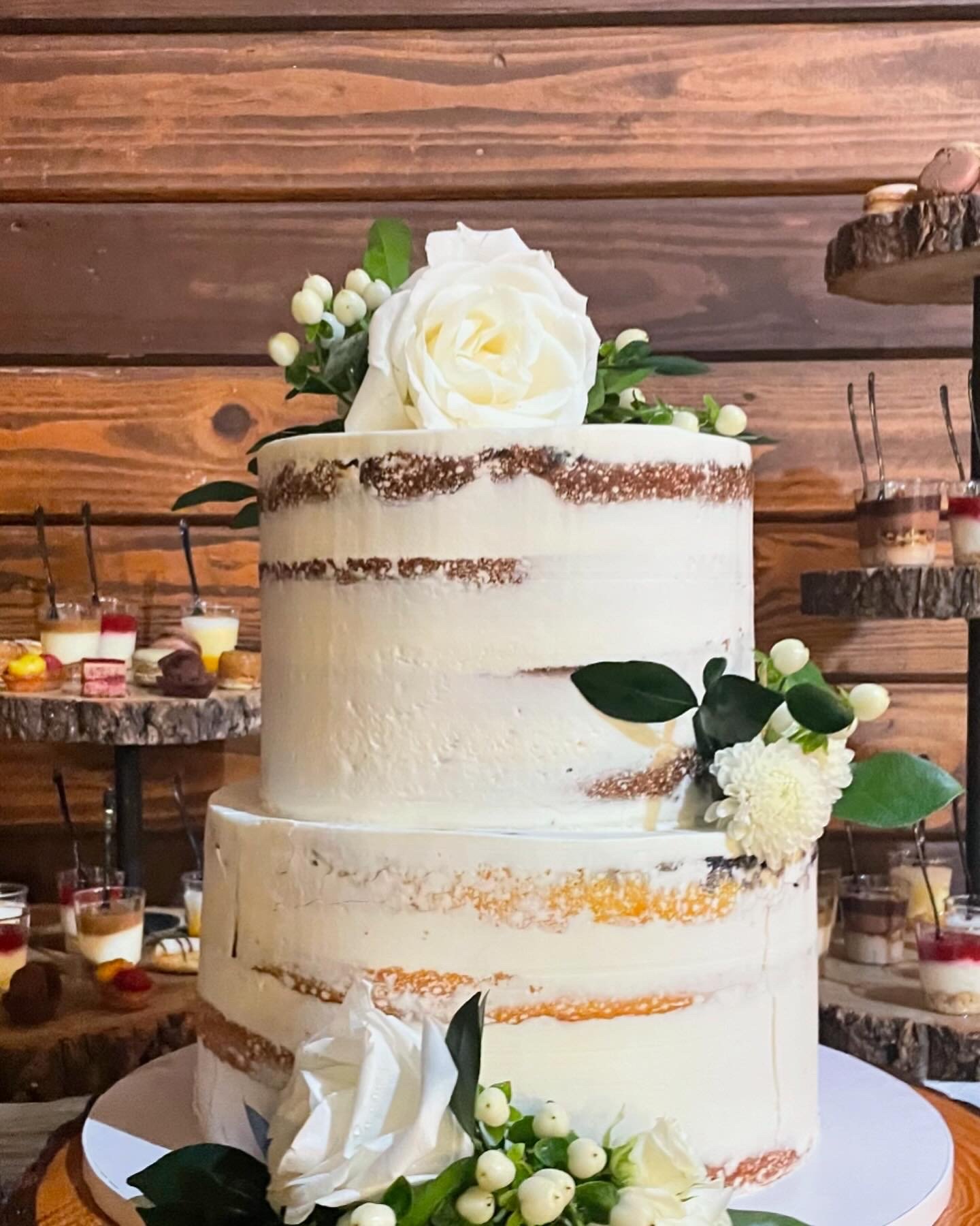 Three-tier vanilla cake decorated with white flowers and green leaves, with a wooden background and a dessert display in the background.