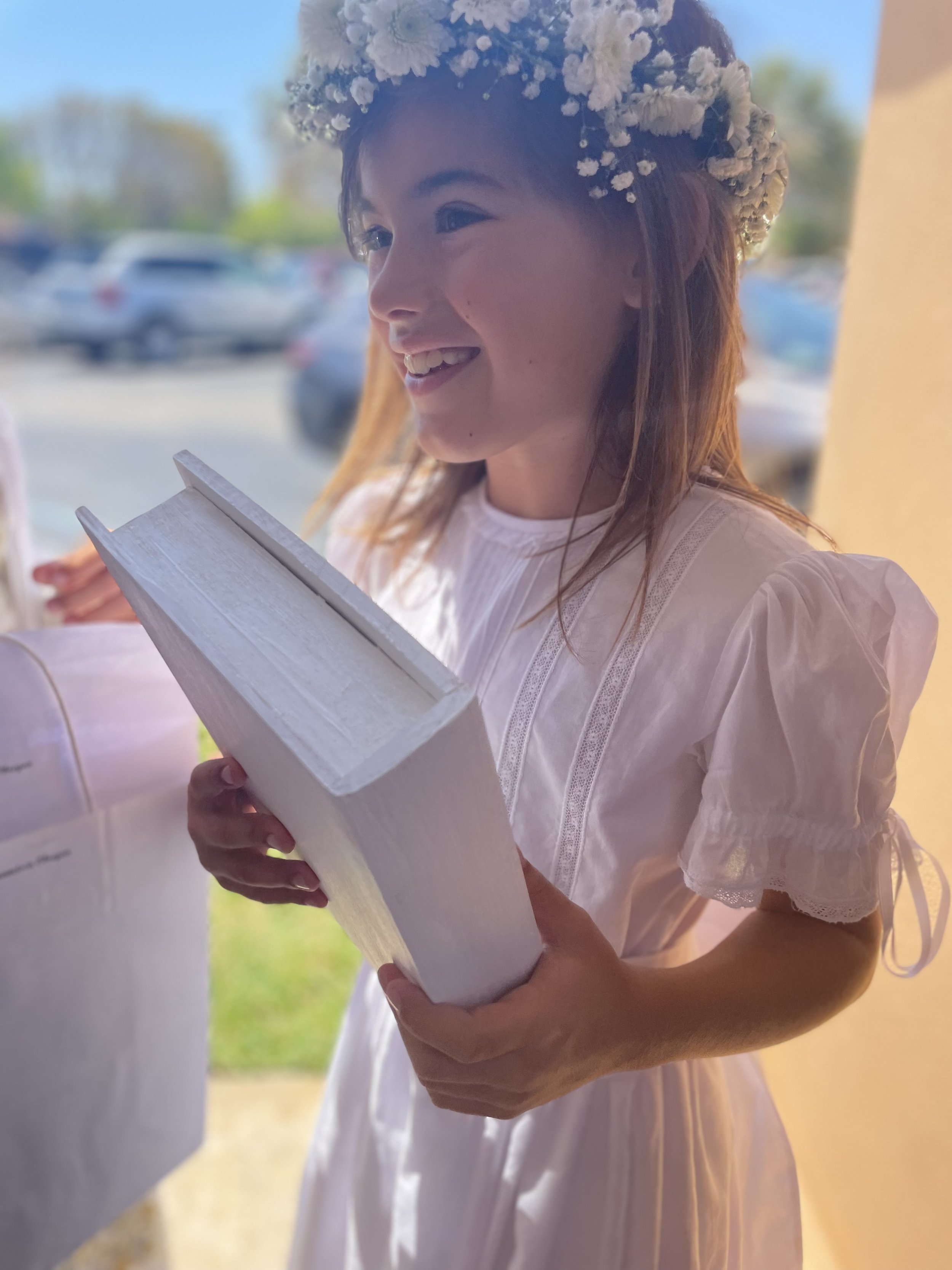 Young girl in a white dress with puffed sleeves and a flower crown, smiling and holding a white book, outdoors on a sunny day.