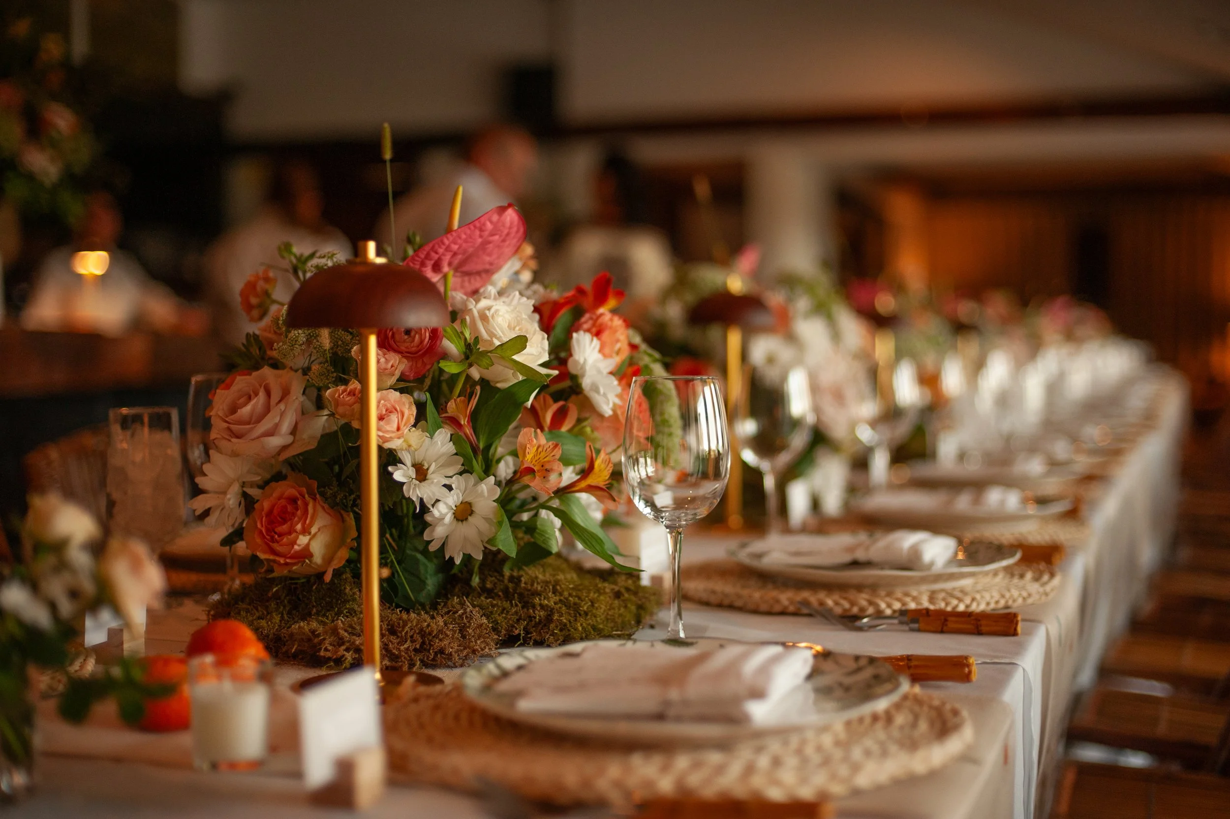 A long dining table decorated with a floral centerpiece of roses, daisies, and other flowers, set with glasses, napkins, and woven placemats for a formal event in a warm-lit room.