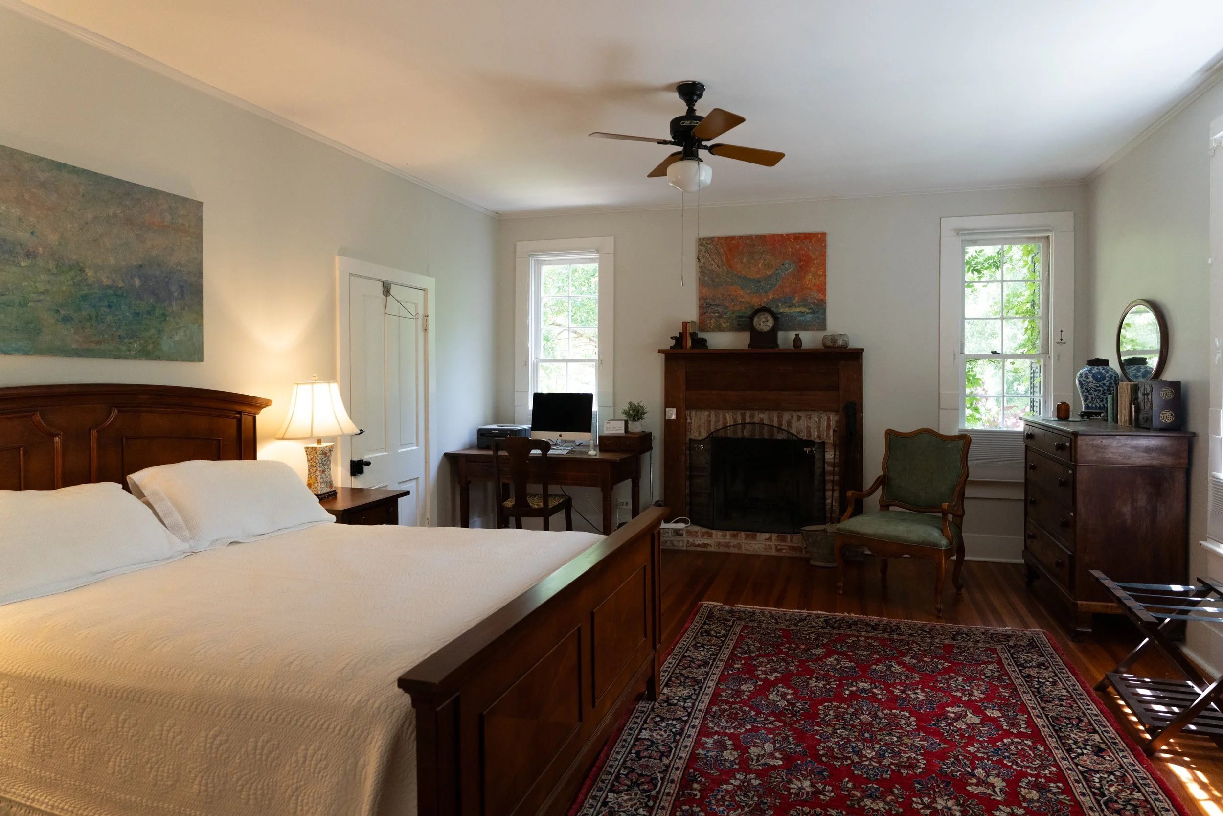 Primary bedroom with queen bed, private bathroom access and a working fireplace. A large picture window sits directly across from the bed with a view of the front gardens. Smaller windows sit on either side of the fireplace. Black out shades.