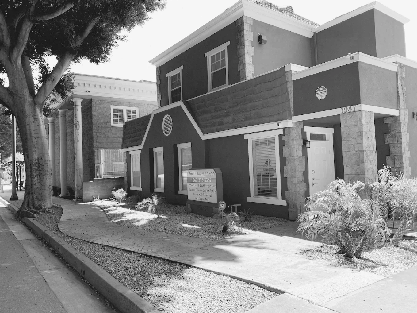 A two-story building with dark exterior walls and stone accents, housing South Bay Med Spa, with a sidewalk, trees, and landscaped plants in front.