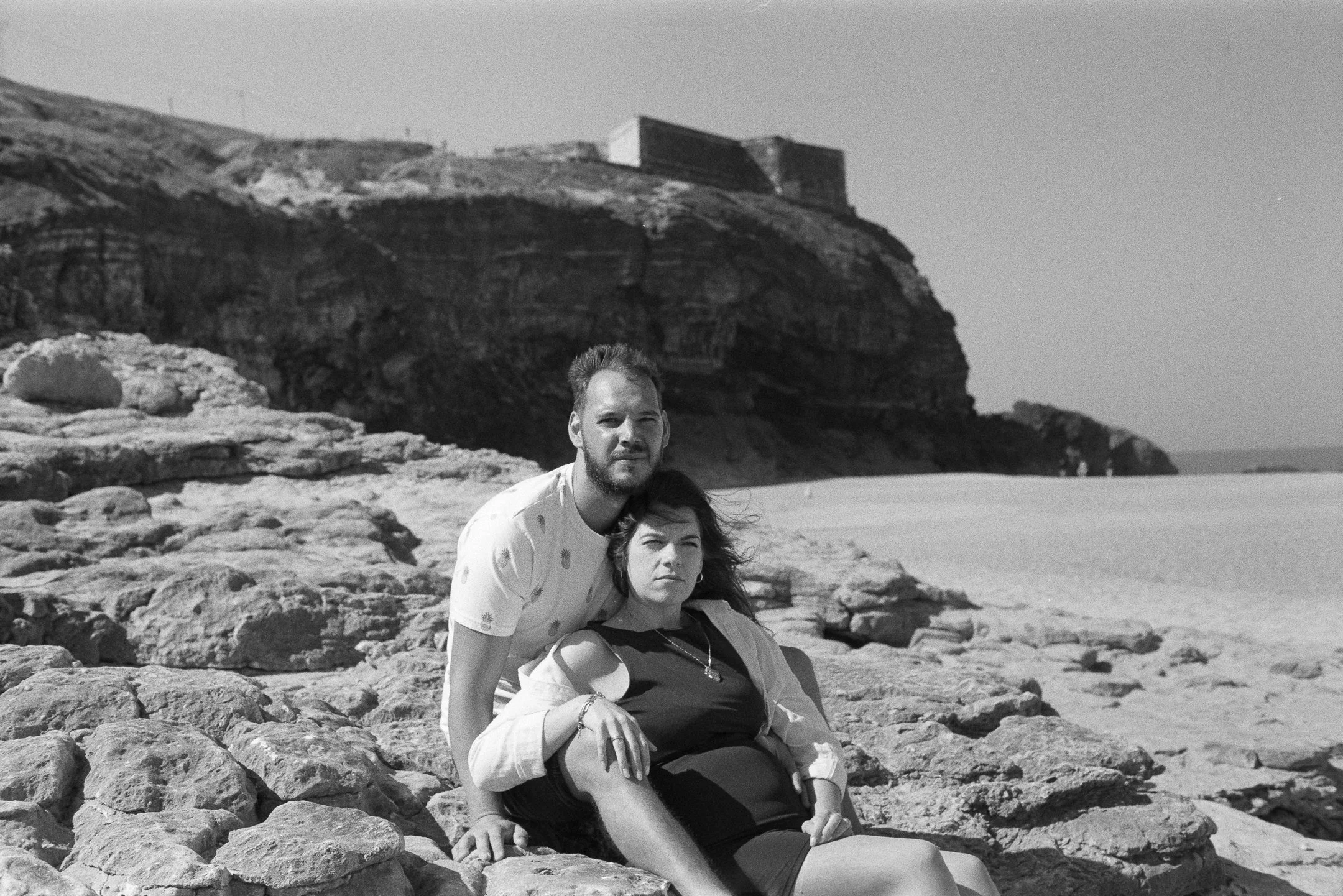 A black and white photo of a man and woman on a rocky beach with cliffs in the background. The man is standing behind the woman, who is sitting on the rocks. The landscape includes sandy shores and a distant shoreline.