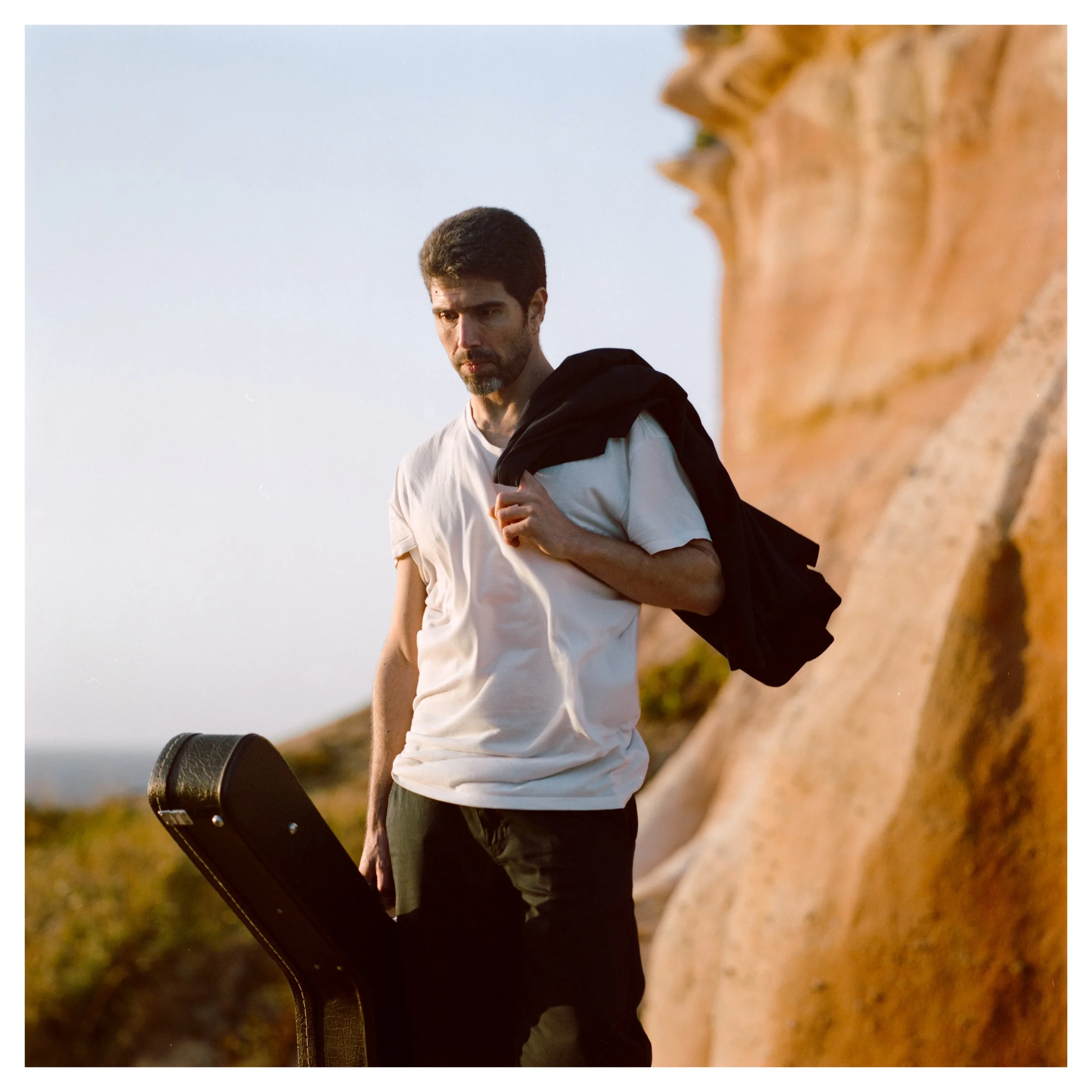 A man with dark hair and a beard stands outdoors near a rocky formation, holding a guitar case and holding a dark jacket over his shoulder, looking pensively downward.