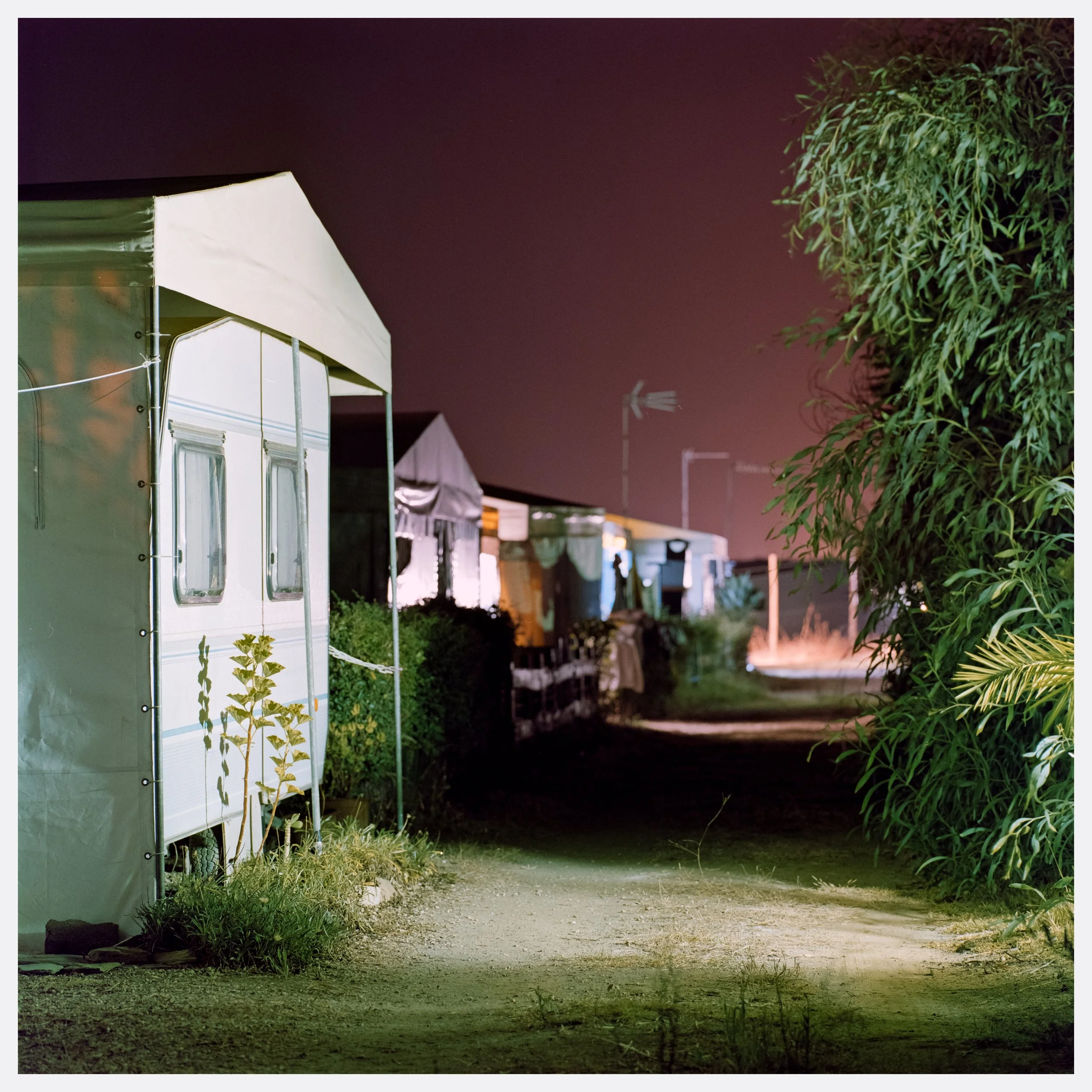 Night view of a row of mobile homes or caravans with lights on, alongside a dirt path bordered by bushes and trees, in a possibly rural or campground setting.