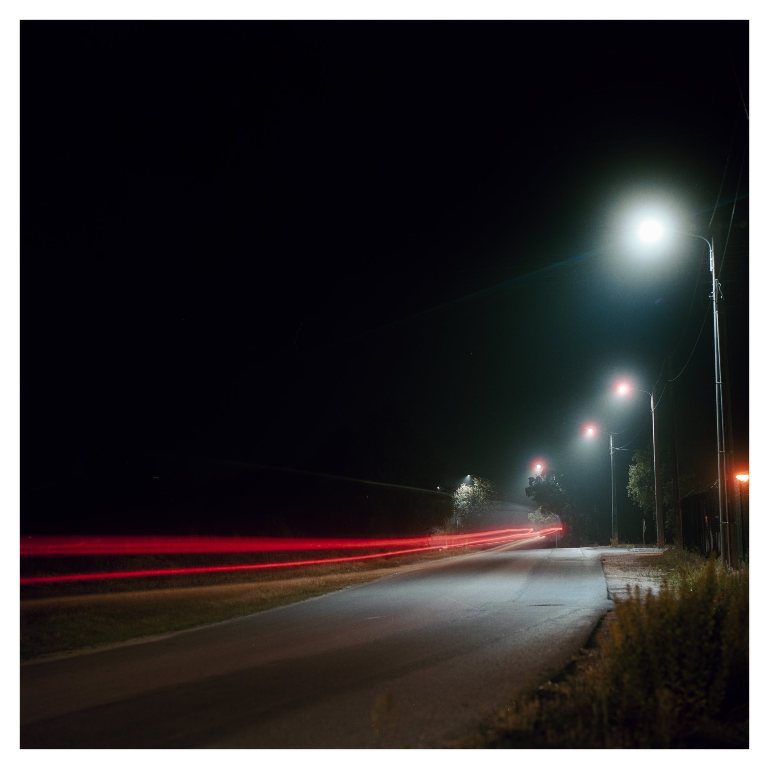 Nighttime street scene with streetlights, a paved road with light trails from passing vehicles, and a dark sky.