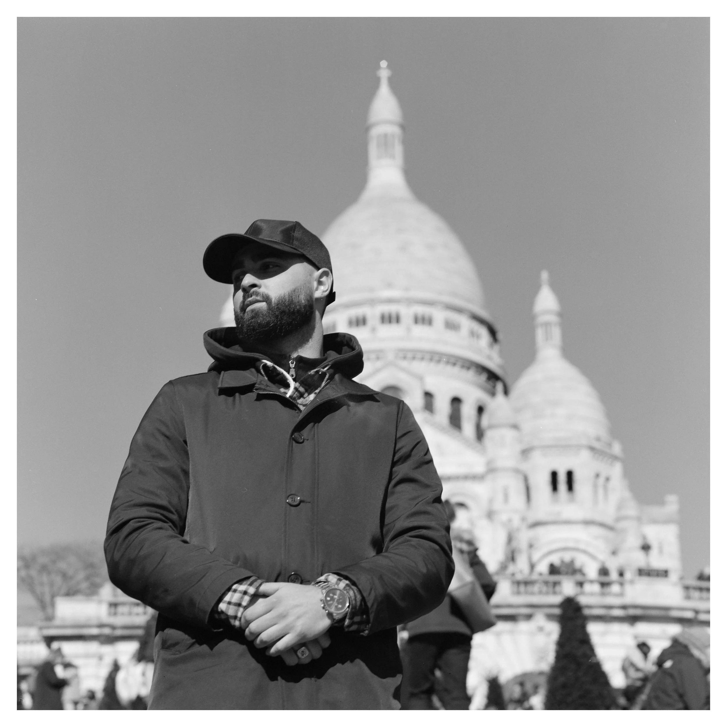 A man with a beard wearing a cap and jacket, standing outdoors with the Sacré-Cœur Basilica in Paris in the background.