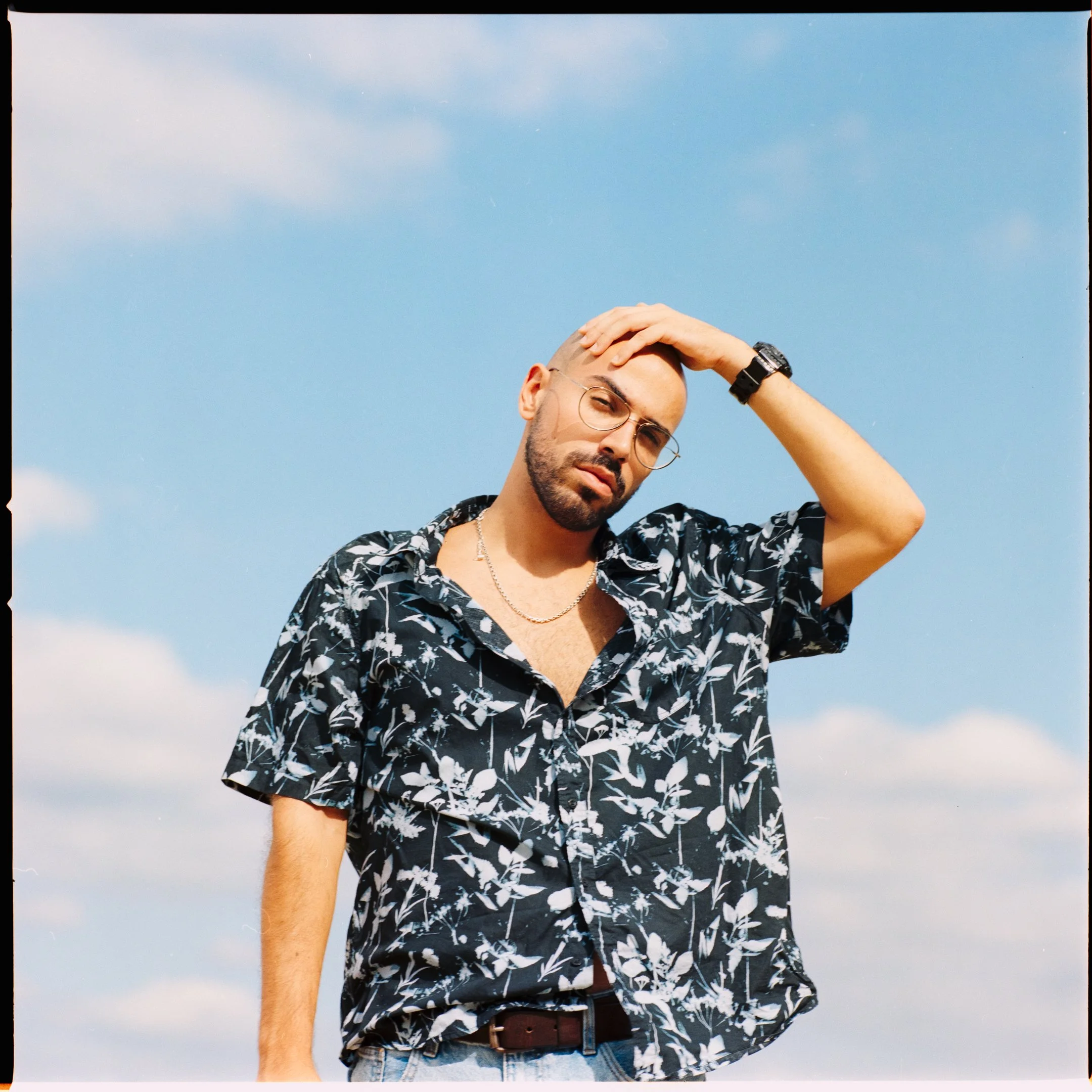 A man with a beard, glasses, and a bald head, touching his forehead with one hand, standing outdoors under a clear blue sky with some clouds, wearing a black short-sleeved shirt with white floral pattern, a silver chain, and a black wristwatch.