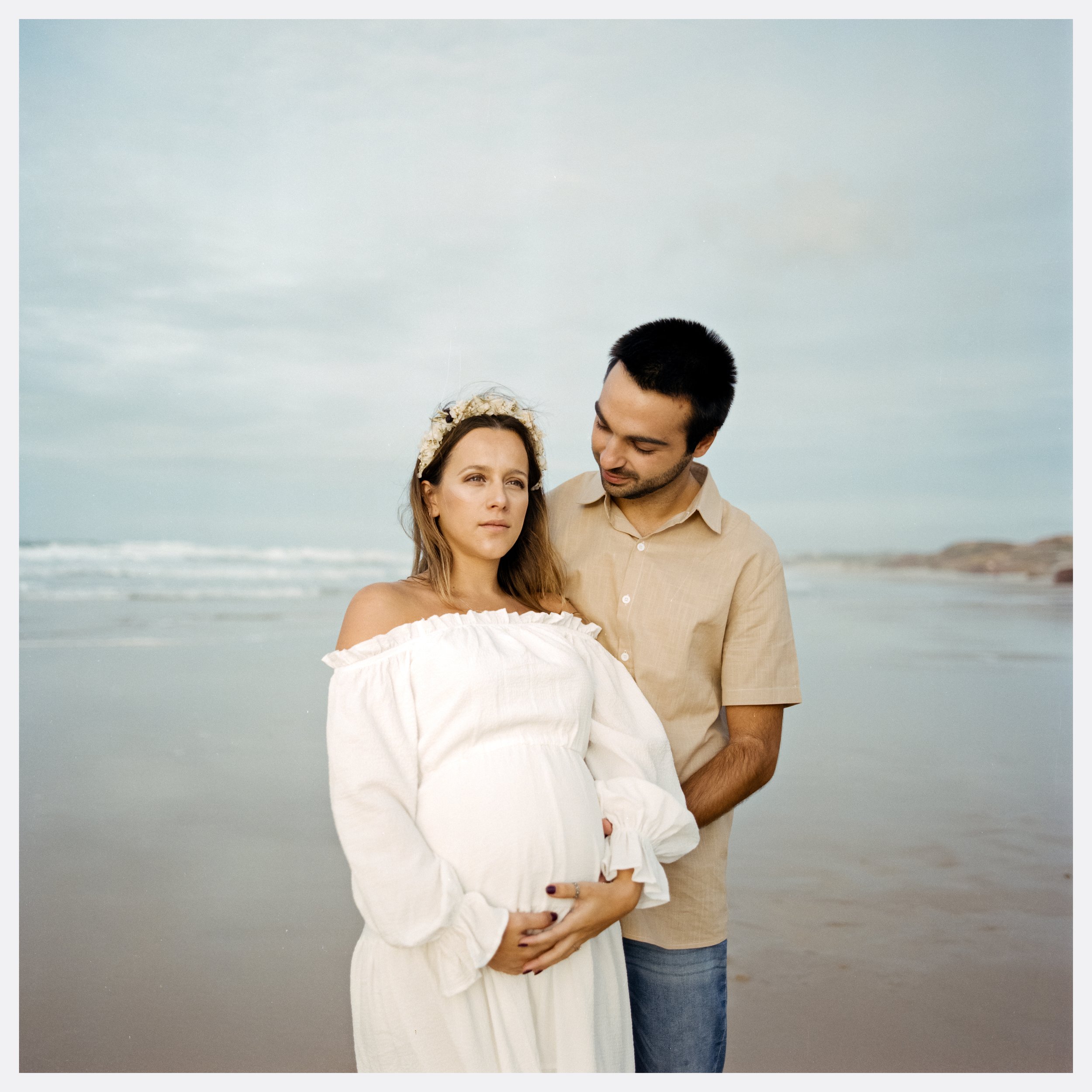 A pregnant woman in a white off-shoulder dress and floral headband standing on the beach, with a man in a beige shirt and jeans standing beside her, holding her belly and looking at her. The sky is cloudy and the ocean is in the background.
