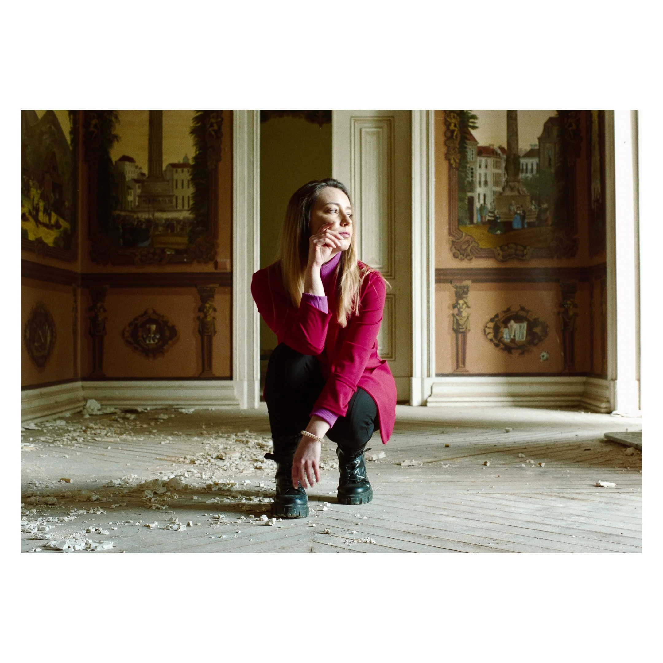 A woman crouching in an abandoned room with peeling floorboards and old painted walls, looking thoughtfully to the side.