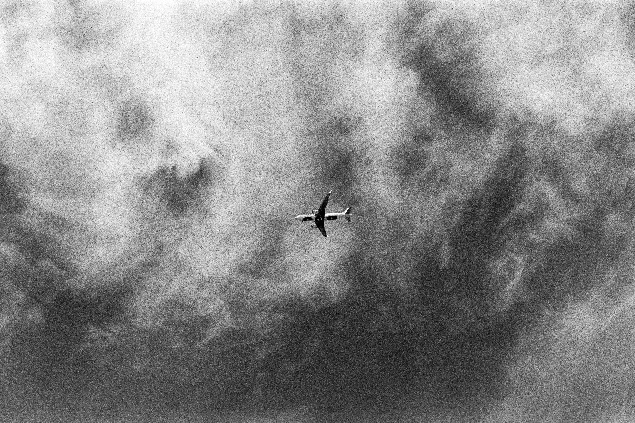 A black and white image of an airplane flying high in the sky amidst clouds. photo by Bruno Novais