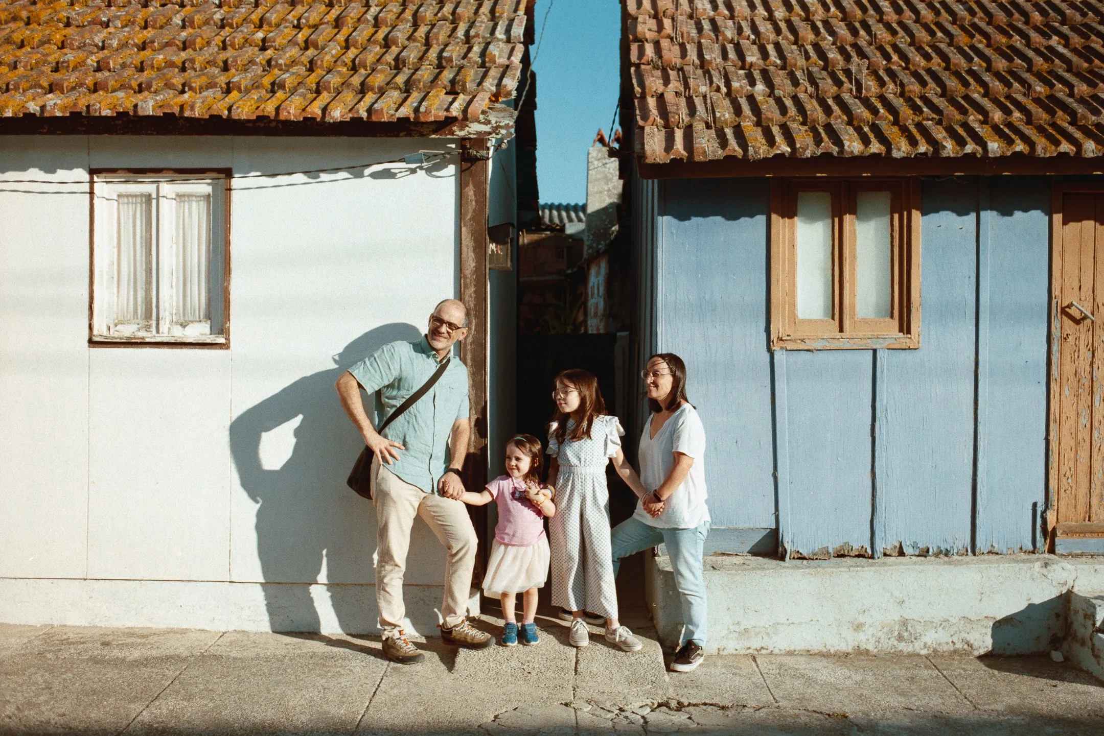 Family of four holding hands and smiling outside their house on a sunny day, with older and new buildings and gray sidewalk. photo by Bruno Novais