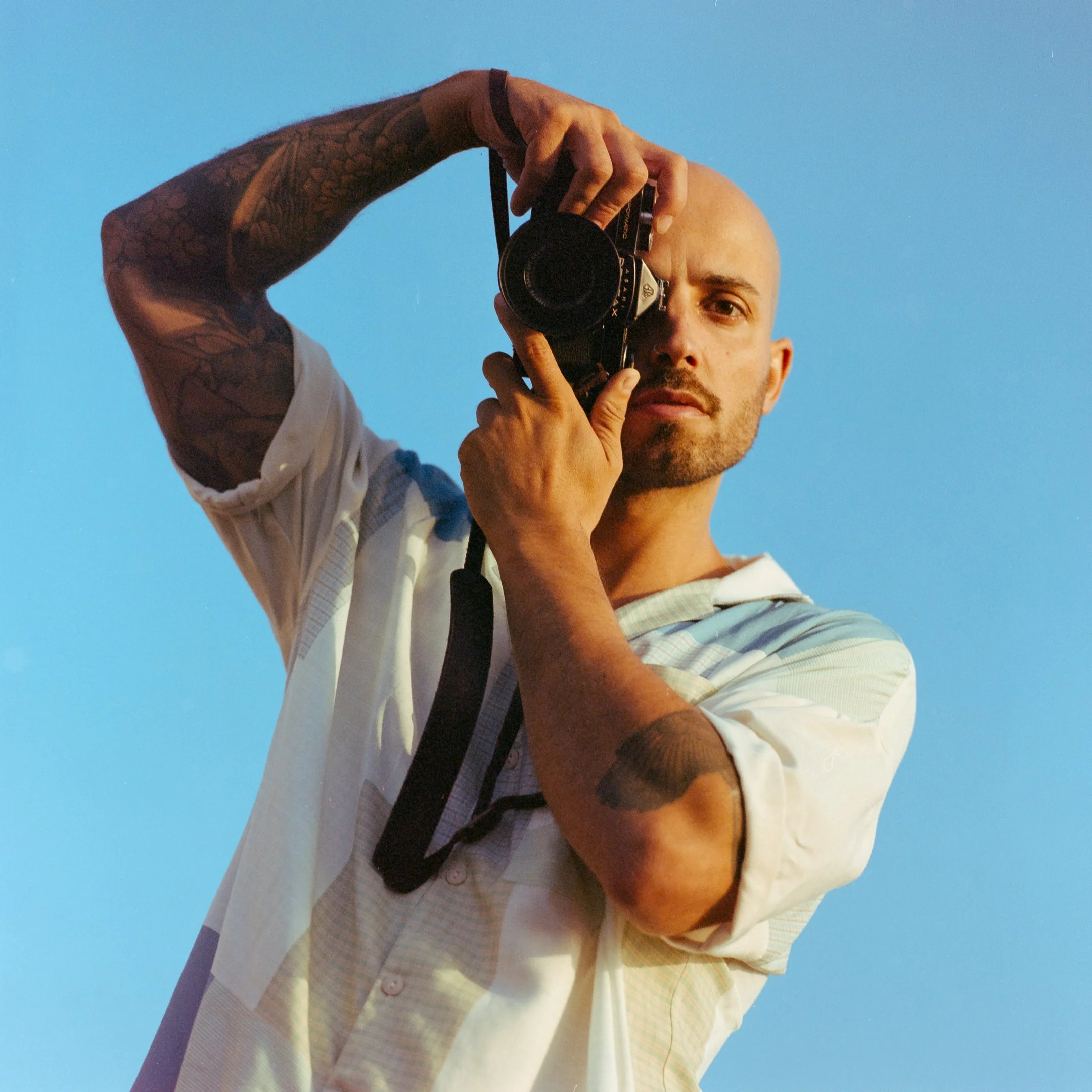 Man taking a photo with a camera against a blue sky, wearing a light-colored short-sleeve shirt and having tattoos on his arms and the side of his face.
