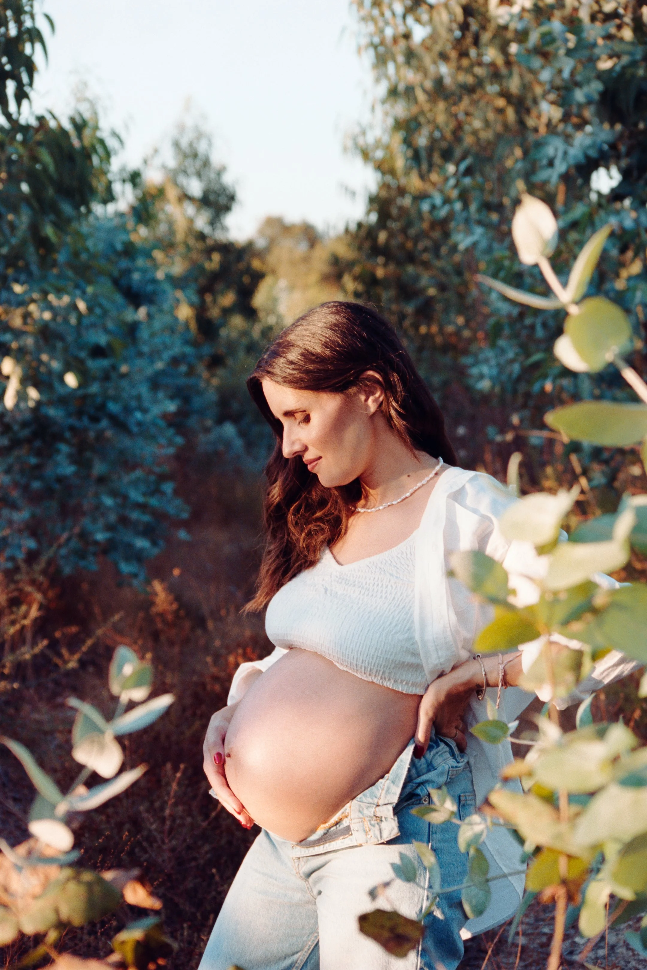 Pregnant woman standing in a garden, touching her belly, with trees and foliage around her during sunset. photo by Bruno Novais