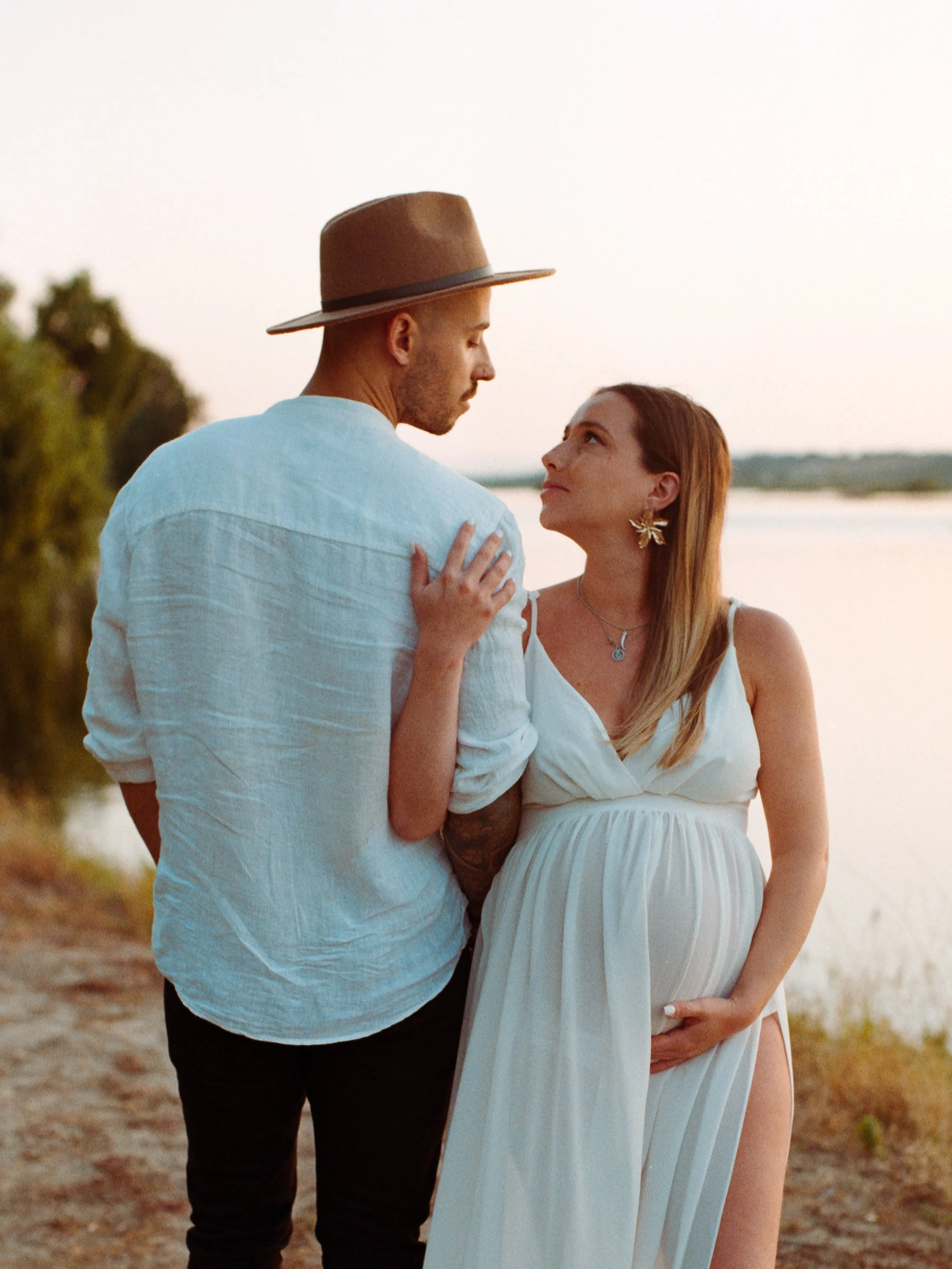 A pregnant woman and a man standing close by a river at sunset, gazing into each other's eyes.