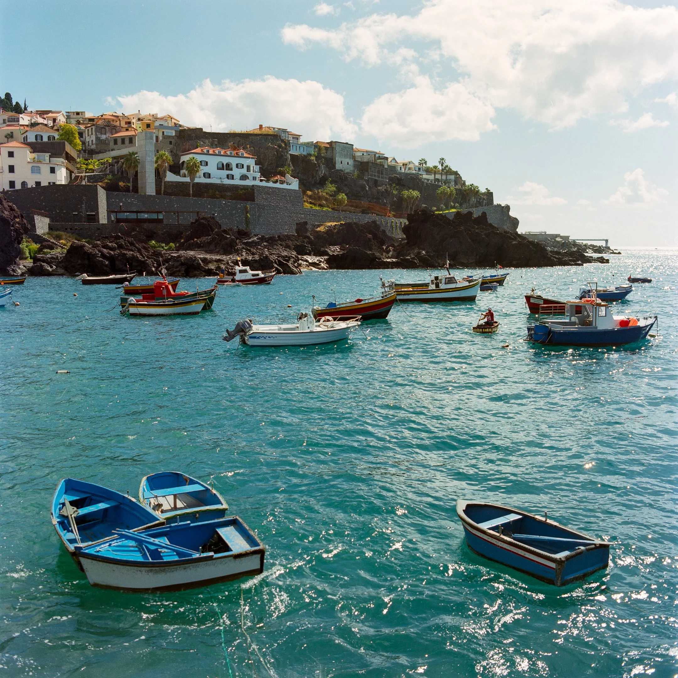 Boats anchored in a clear blue bay with a hillside town featuring white houses with red and orange roofs in the background, under a partly cloudy sky.