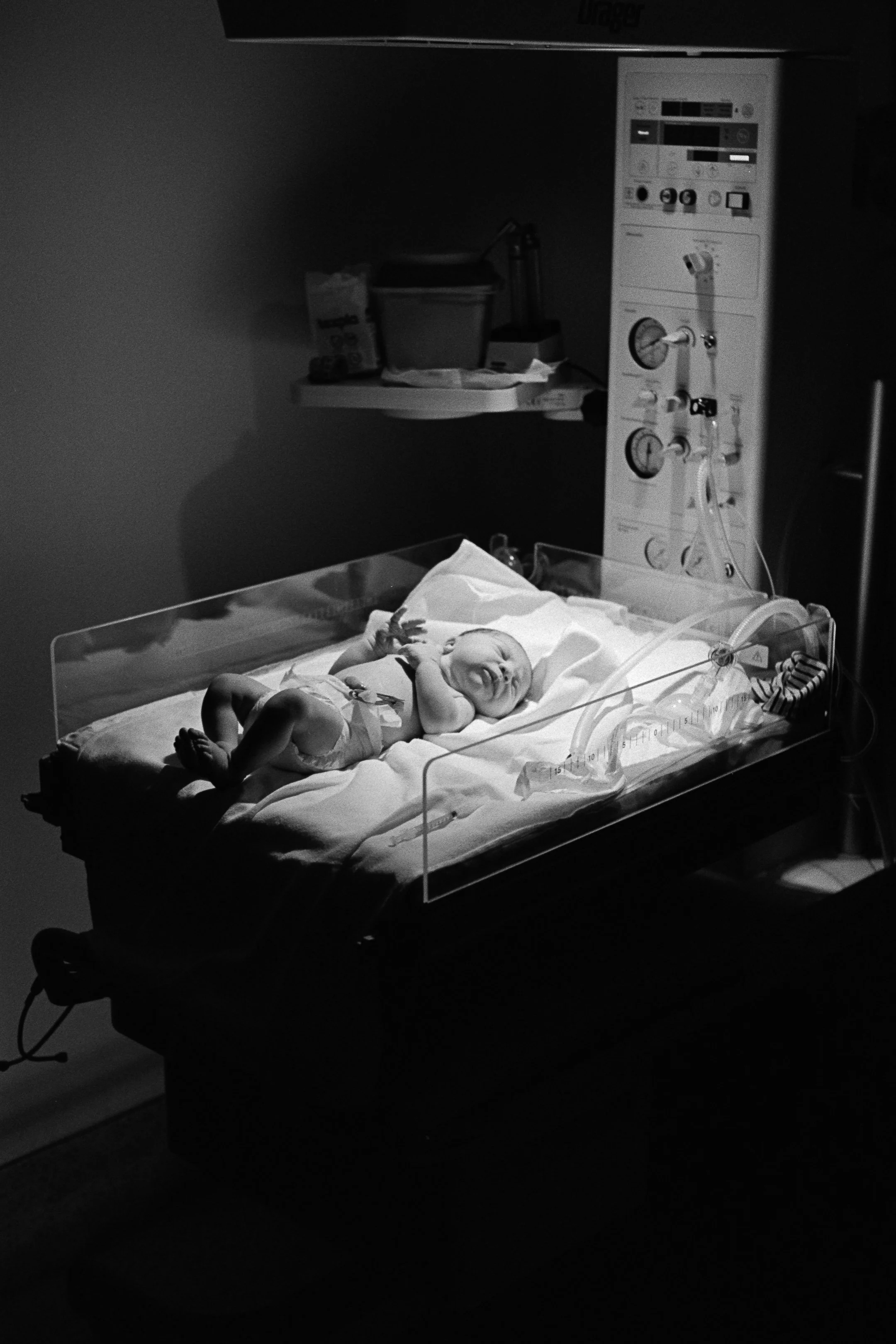 A newborn baby lying in a clear hospital bassinet, smiling and holding its hands near its face, in a hospital room with medical equipment in the background. photo by Bruno Novais