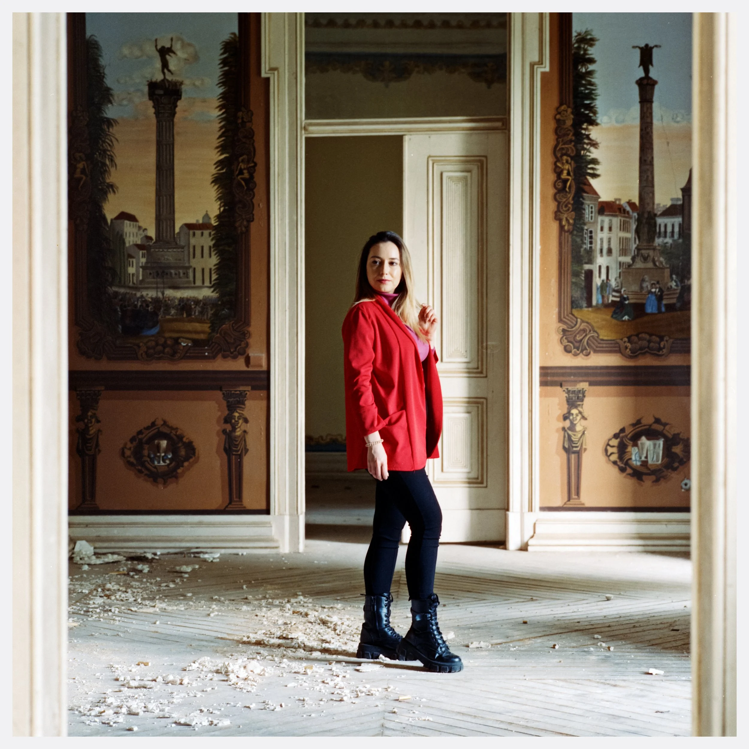 A woman with blonde hair wearing a red jacket, black pants, and black boots standing in a damaged room with debris on the floor and decorative painted walls. photo by Bruno Novais