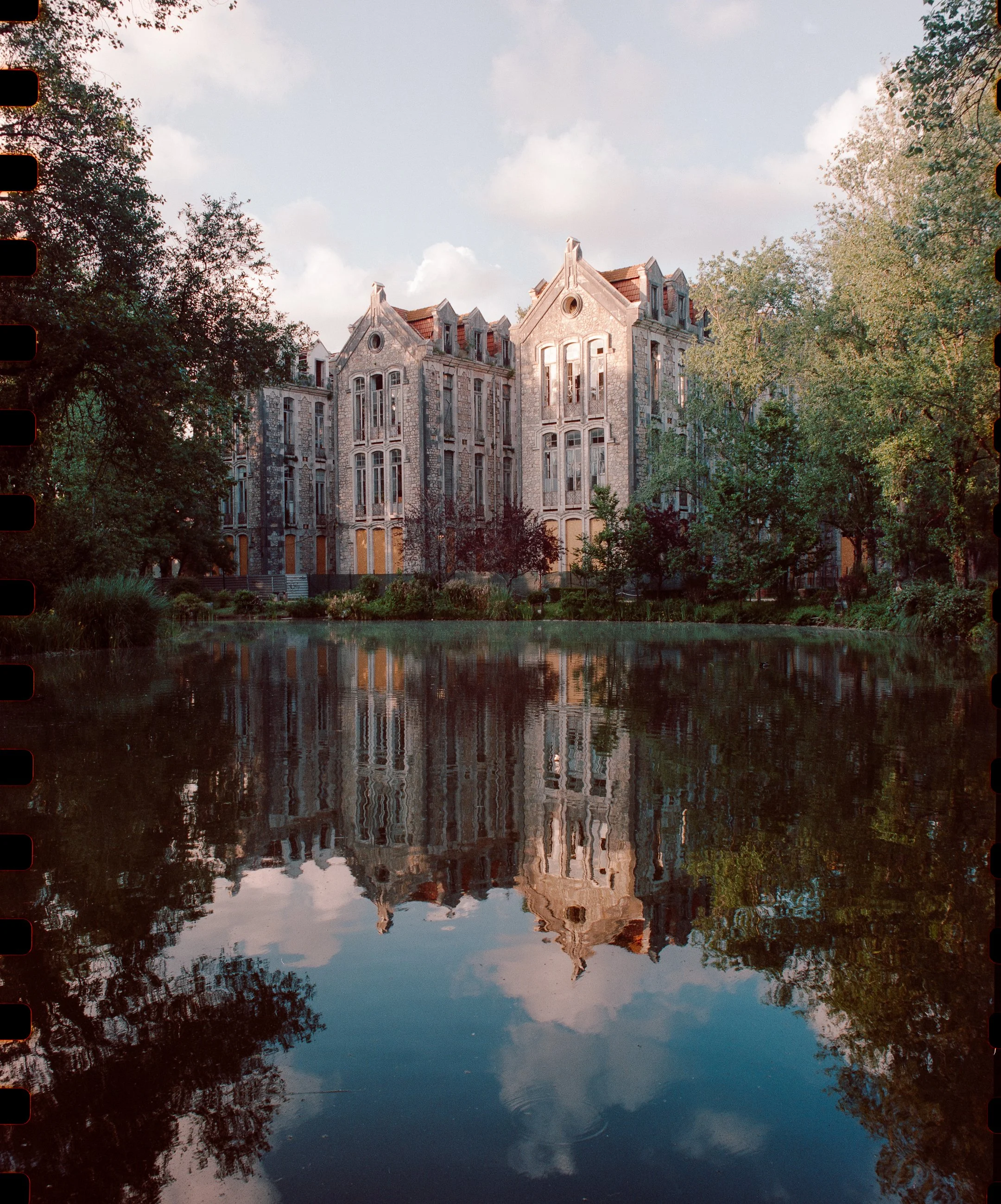 A large, historic stone mansion with multiple peaked gables, tall windows, and red roof sections, situated near a tranquil body of water that reflects the building and surrounding trees.