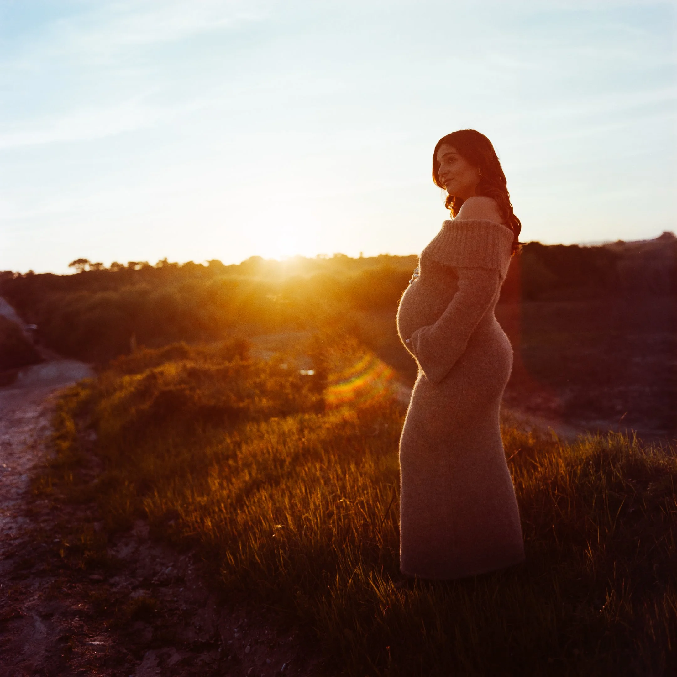 A pregnant woman in a long dress standing outdoors at sunset, with a dirt path and grassy hills in the background.