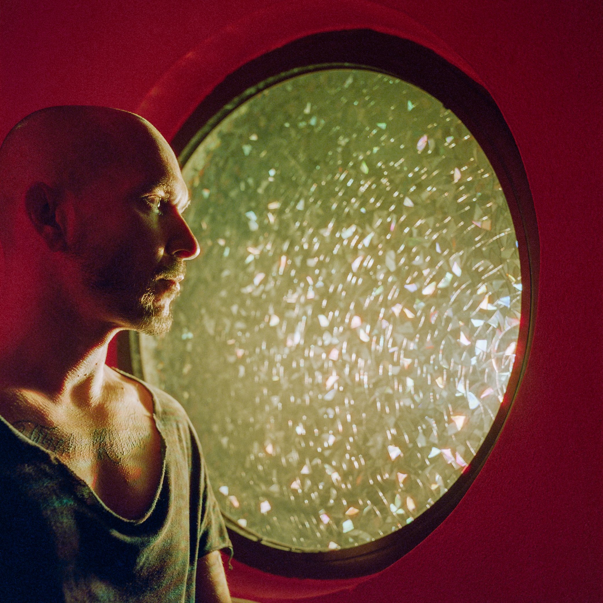 A man with a beard and a tattoo on his shoulder looks out of a round window with a shattered or frosted glass, illuminated by greenish light, in a red-walled room.