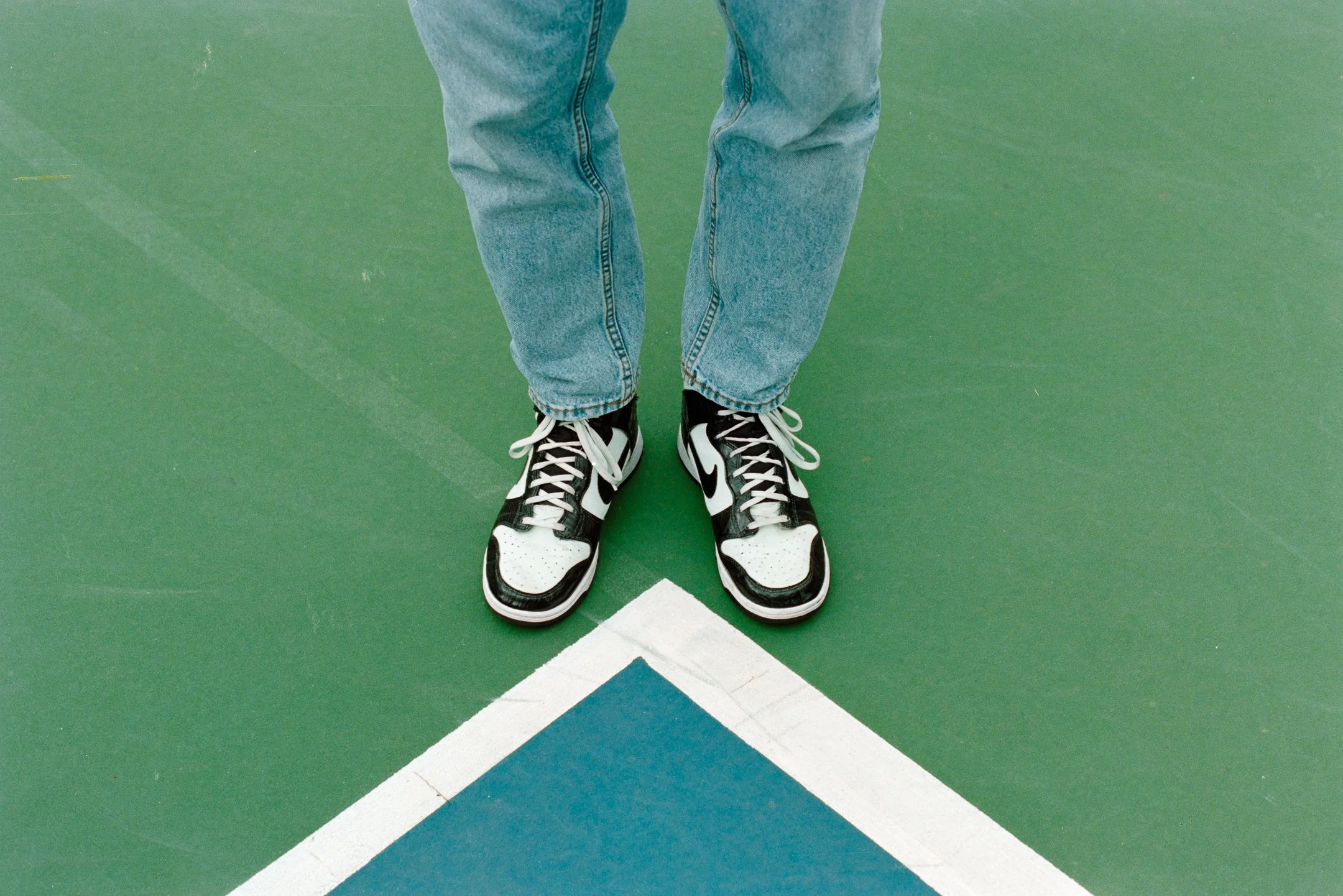 Person standing on a green tennis or basketball court with the tips of their shoes crossing a white and blue court line. photo by Bruno Novais