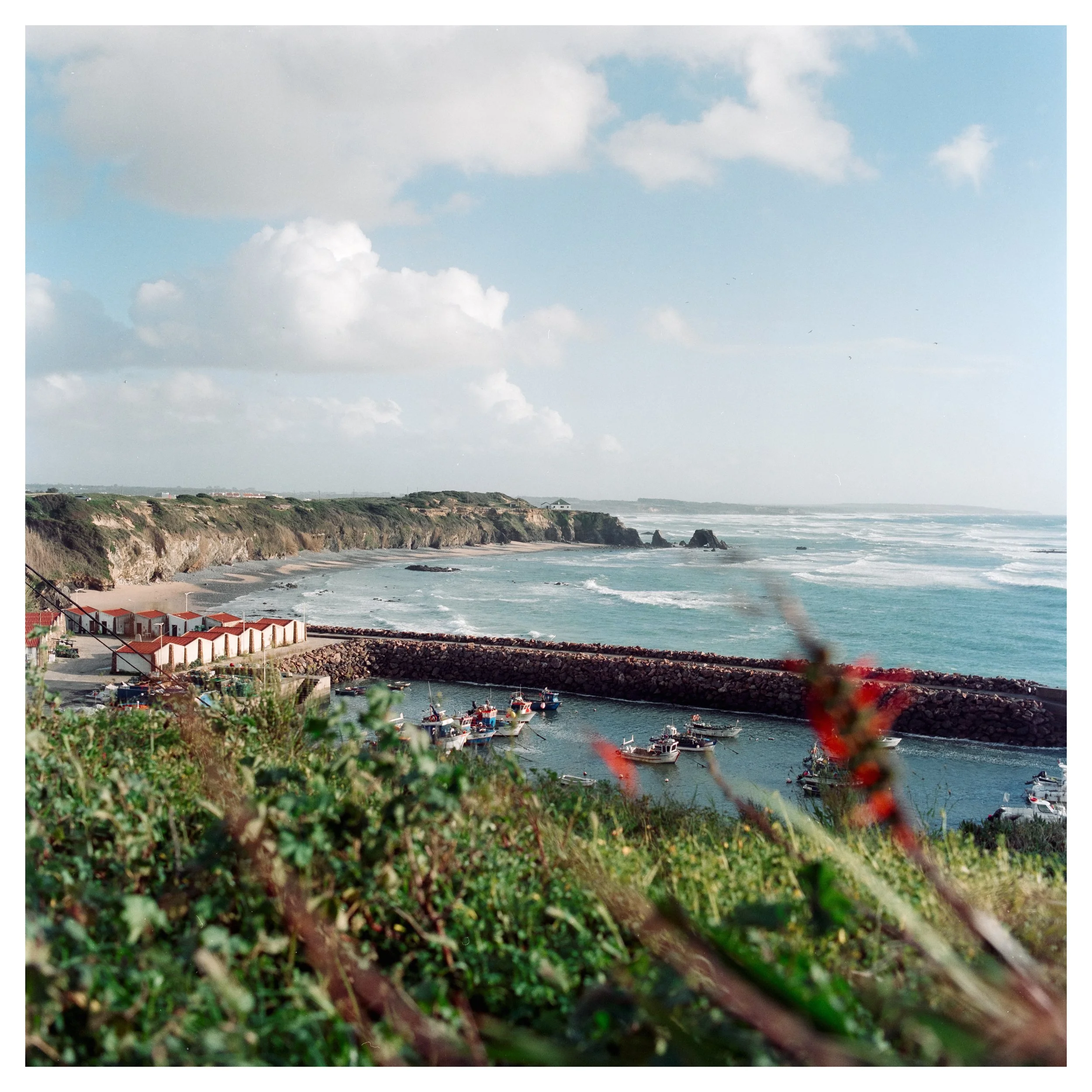 A coastal scene with a harbor and boats near a rocky breakwater, sandy beach, cliffs, and a partly cloudy sky.