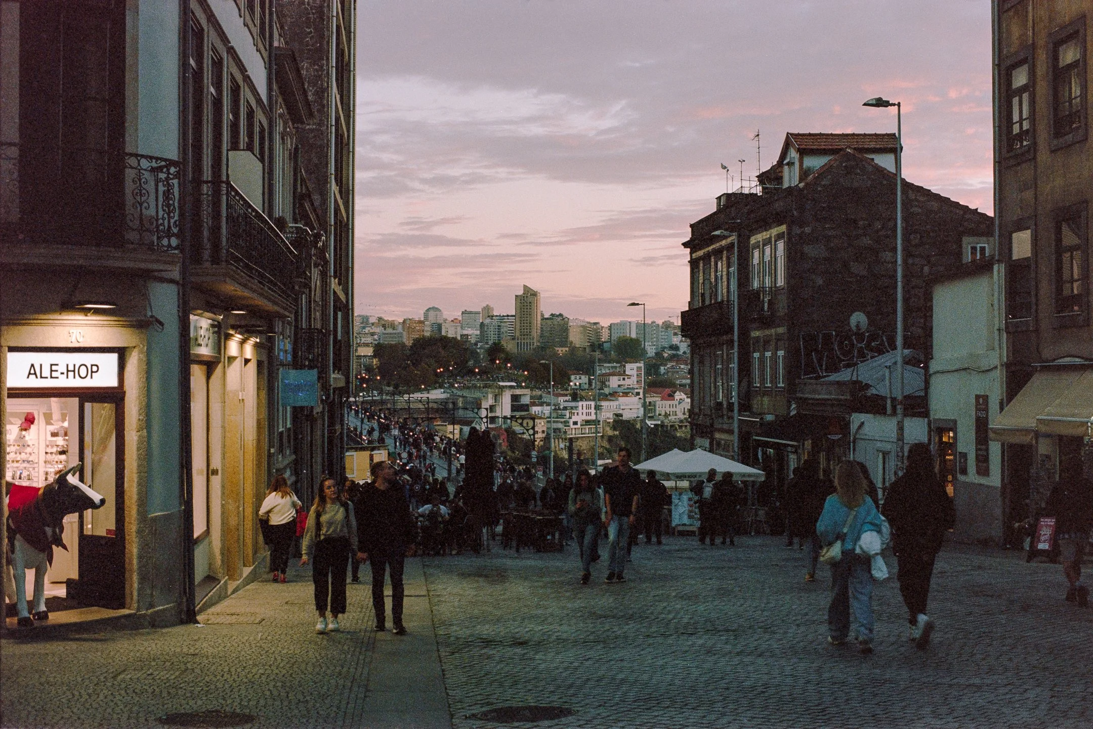 People walking on a cobblestone street at dusk in an urban area with buildings and city skyline in the background. photo by Bruno Novais