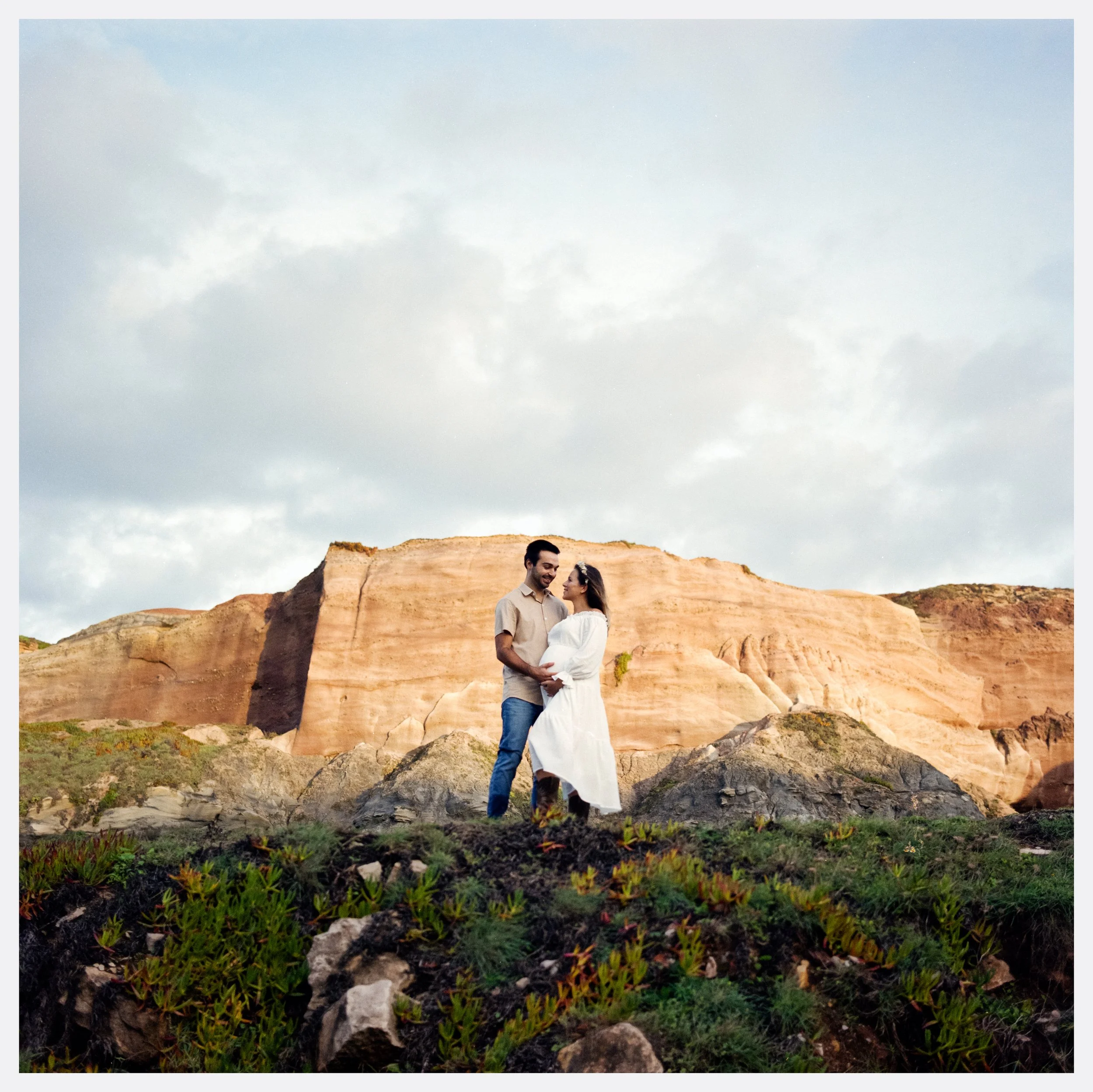 A couple standing on rocks in a scenic outdoor landscape with large sandstone formations in the background, under a partly cloudy sky.