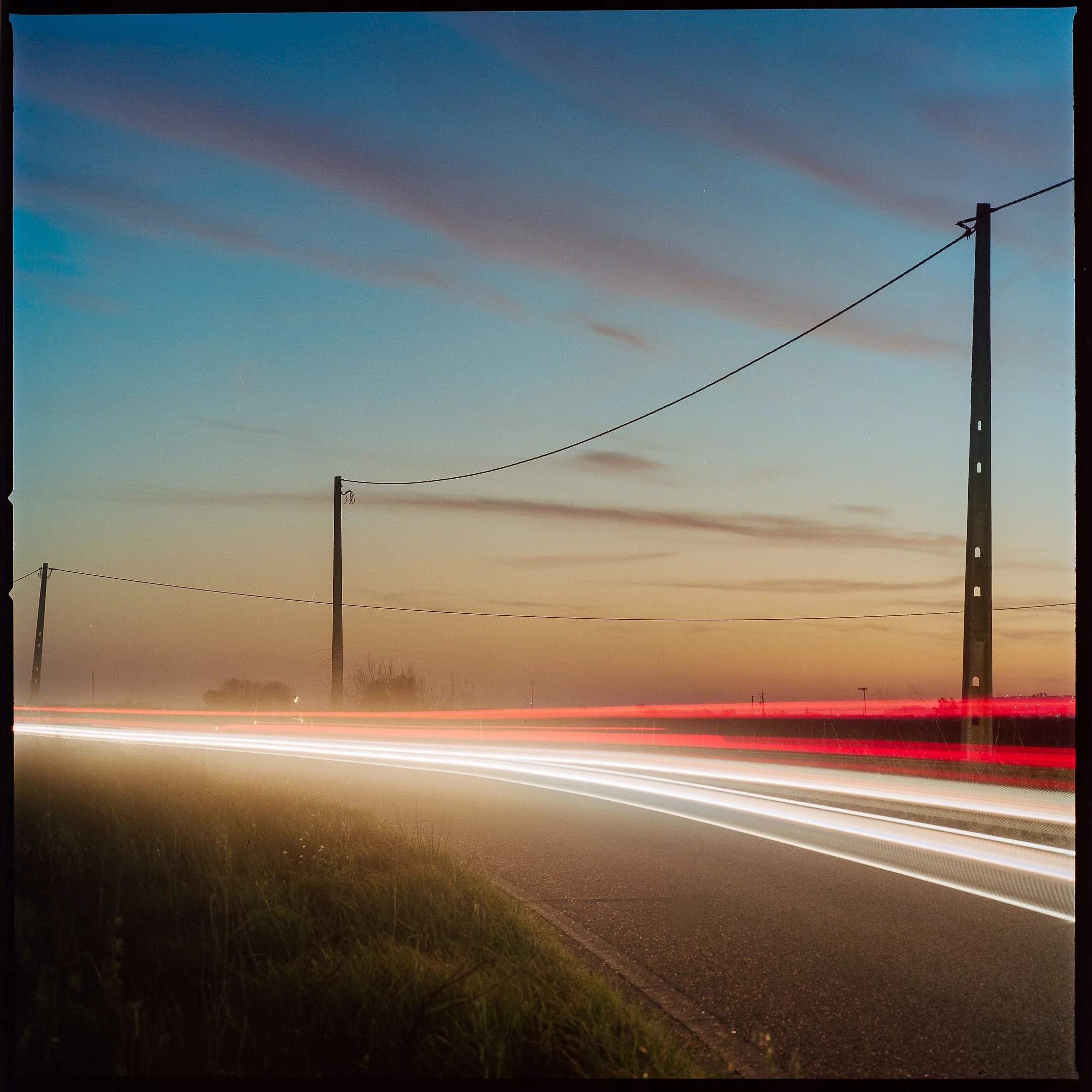 Long exposure photograph of a rural road at sunset with light trails from passing vehicles, power poles, and a colorful sky with a gradient of blue to orange.