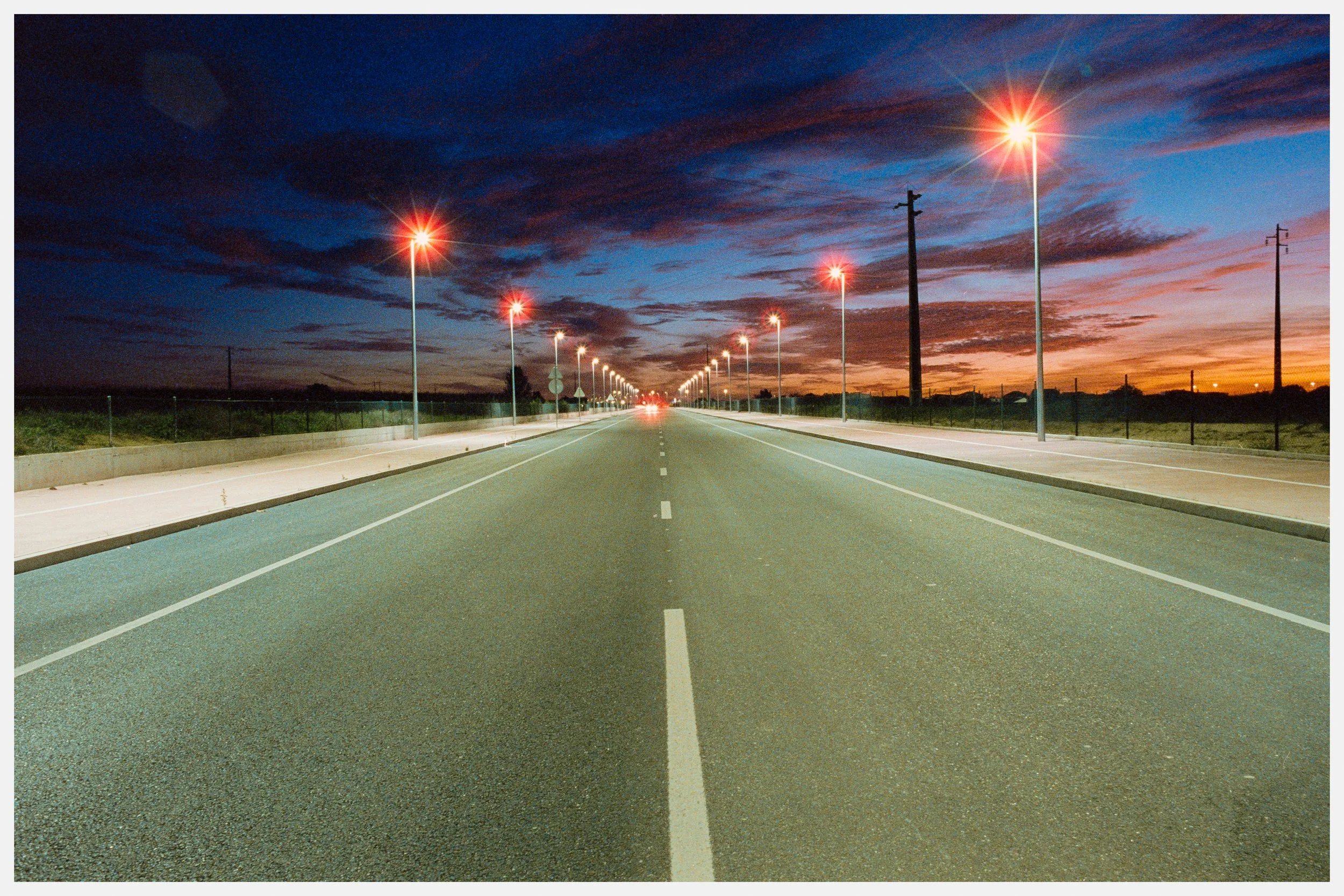 Empty road with streetlights at sunset, dark clouds in the sky, and power lines along the roadside.