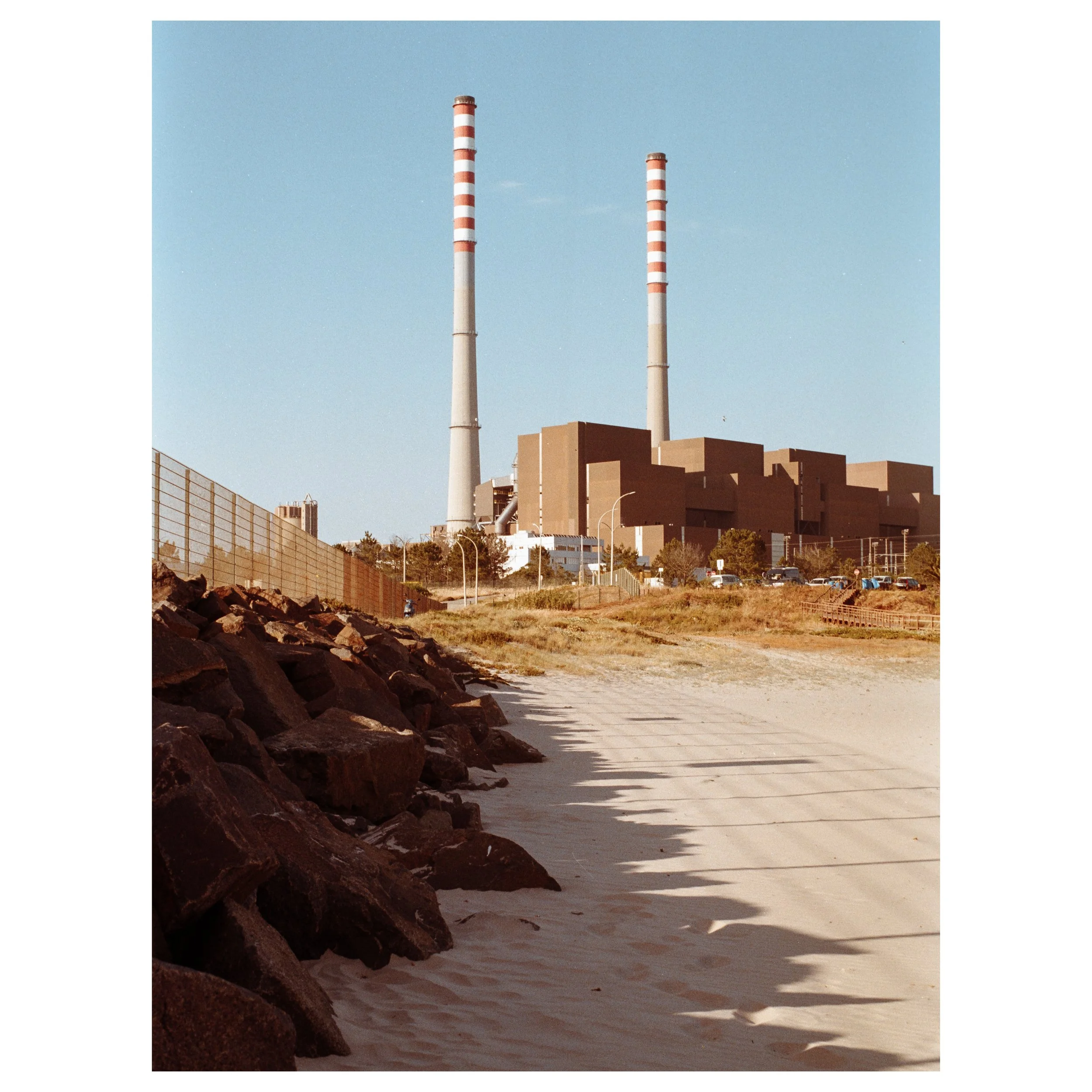 A power plant with two tall smokestacks in a landscape with sandy beach, rocks, and some trees