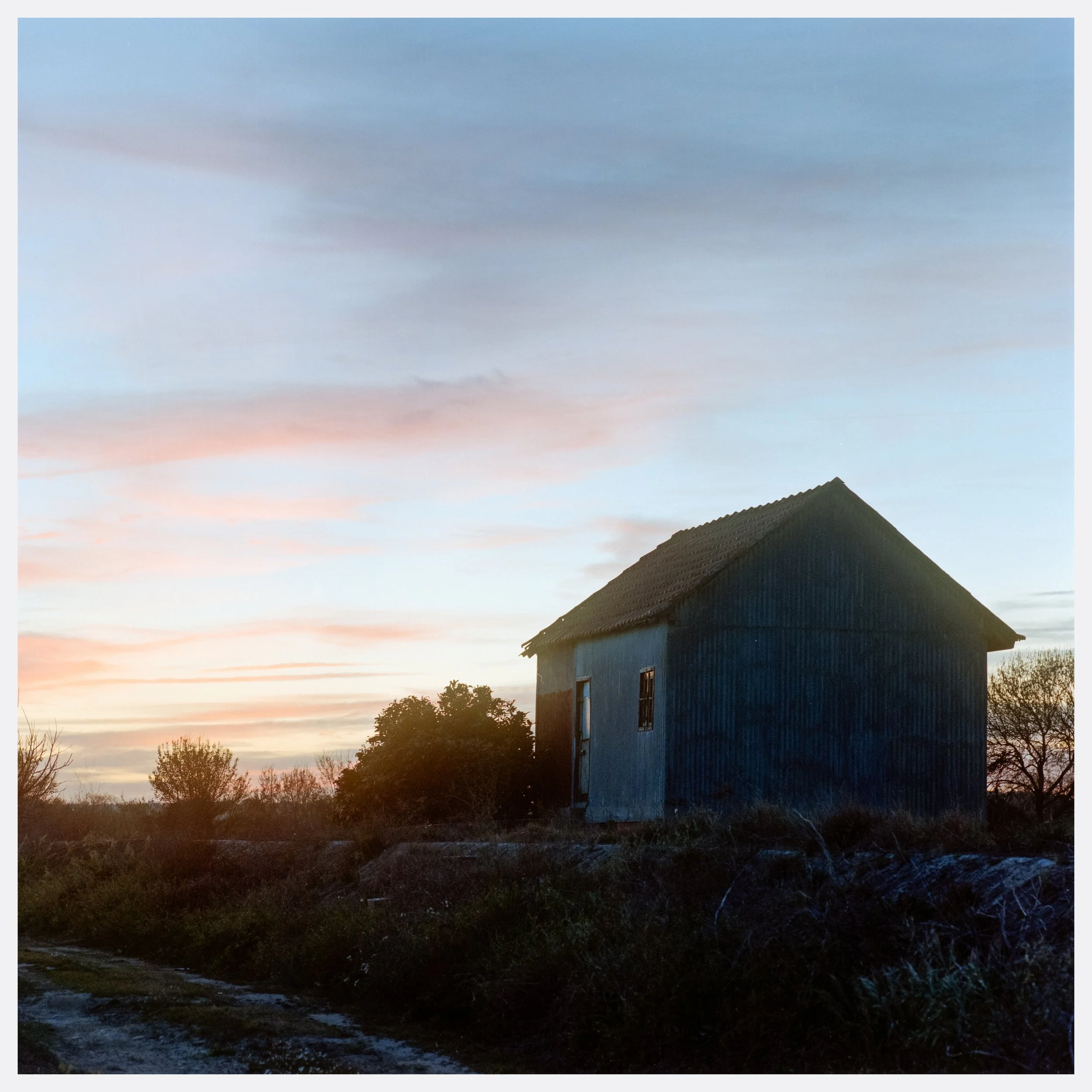 A small, old blue house with a pitched roof stands alone on a field during sunset, surrounded by trees and bushes.