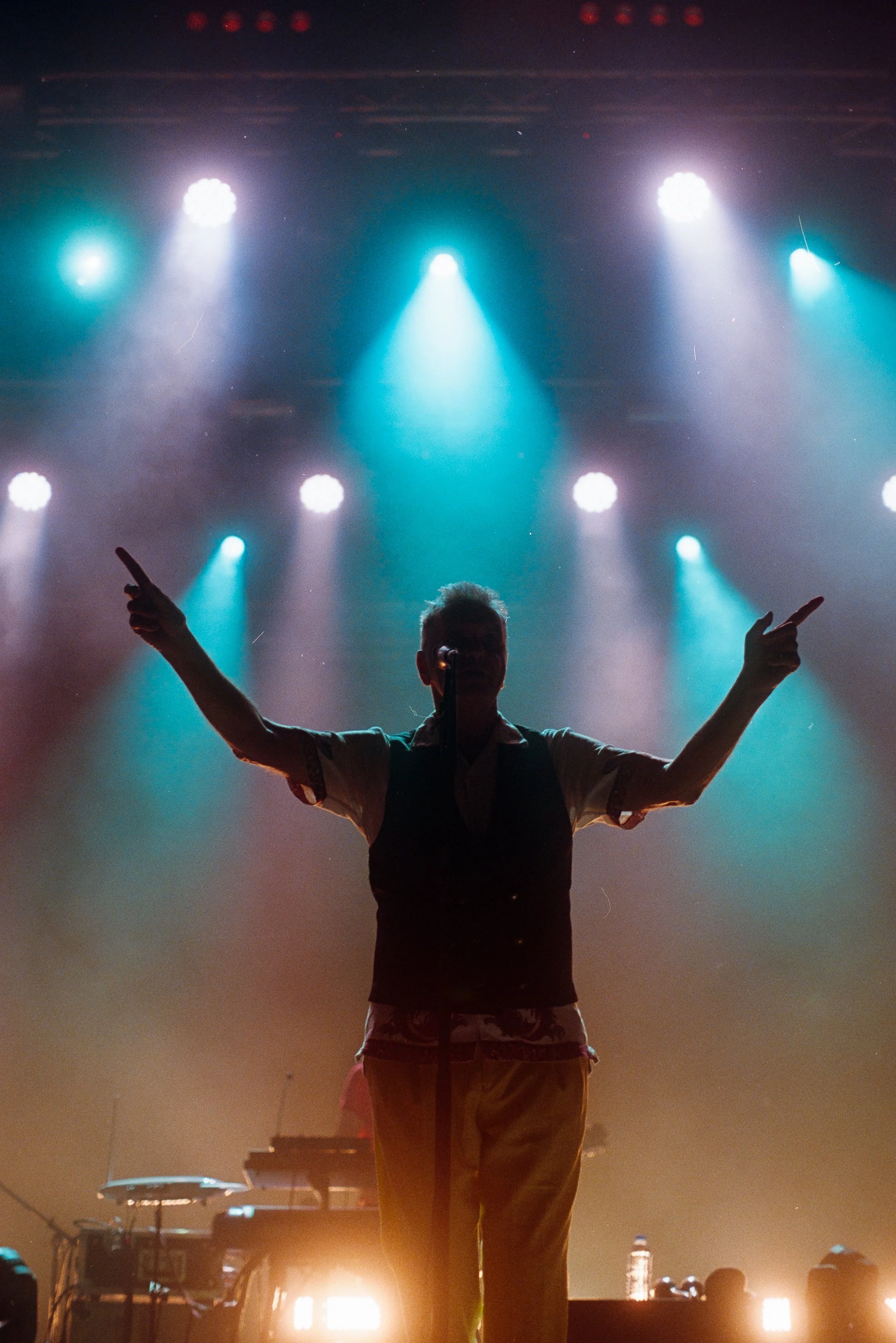 A silhouette of a performer with arms raised in a concert setting with blue and teal stage lights overhead. photo by Bruno Novais
