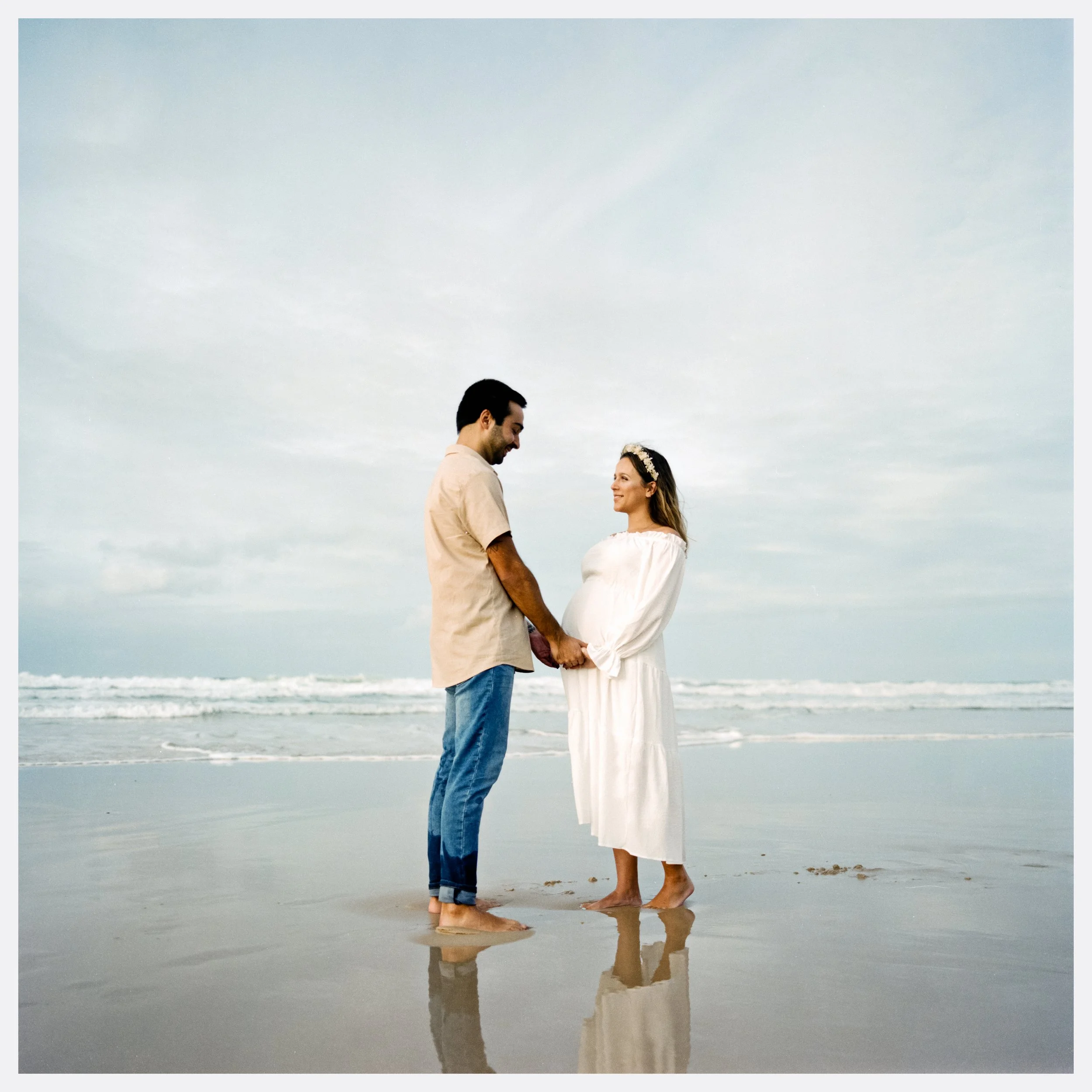 A pregnant woman and a man holding hands on a beach with waves in the background, during daytime.