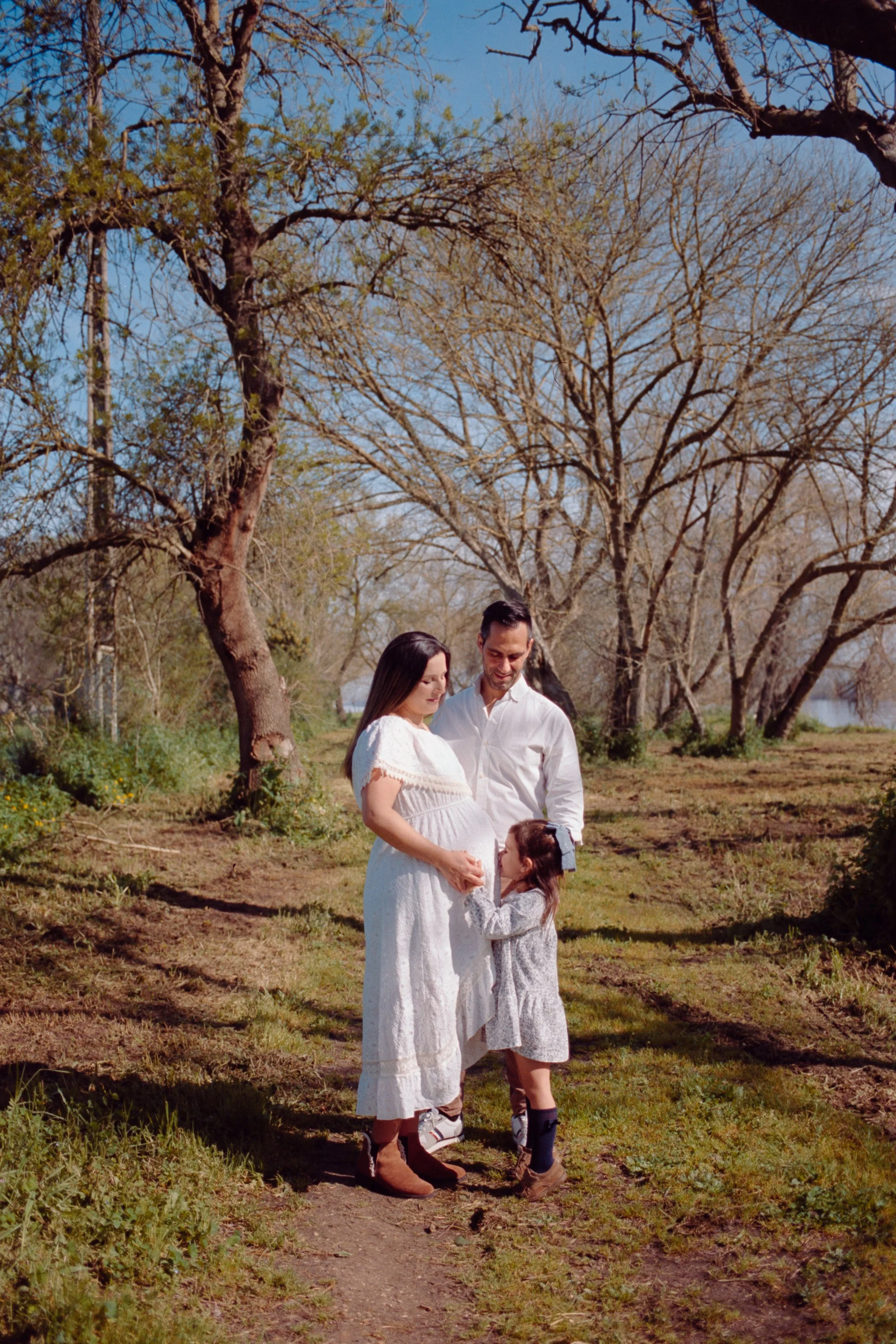 A family of three, pregnant woman, man, and young girl, standing on a dirt path surrounded by trees with bare branches in early spring or late fall. The woman is wearing a white dress, the man in a white shirt, and the girl in a gray dress. They are 