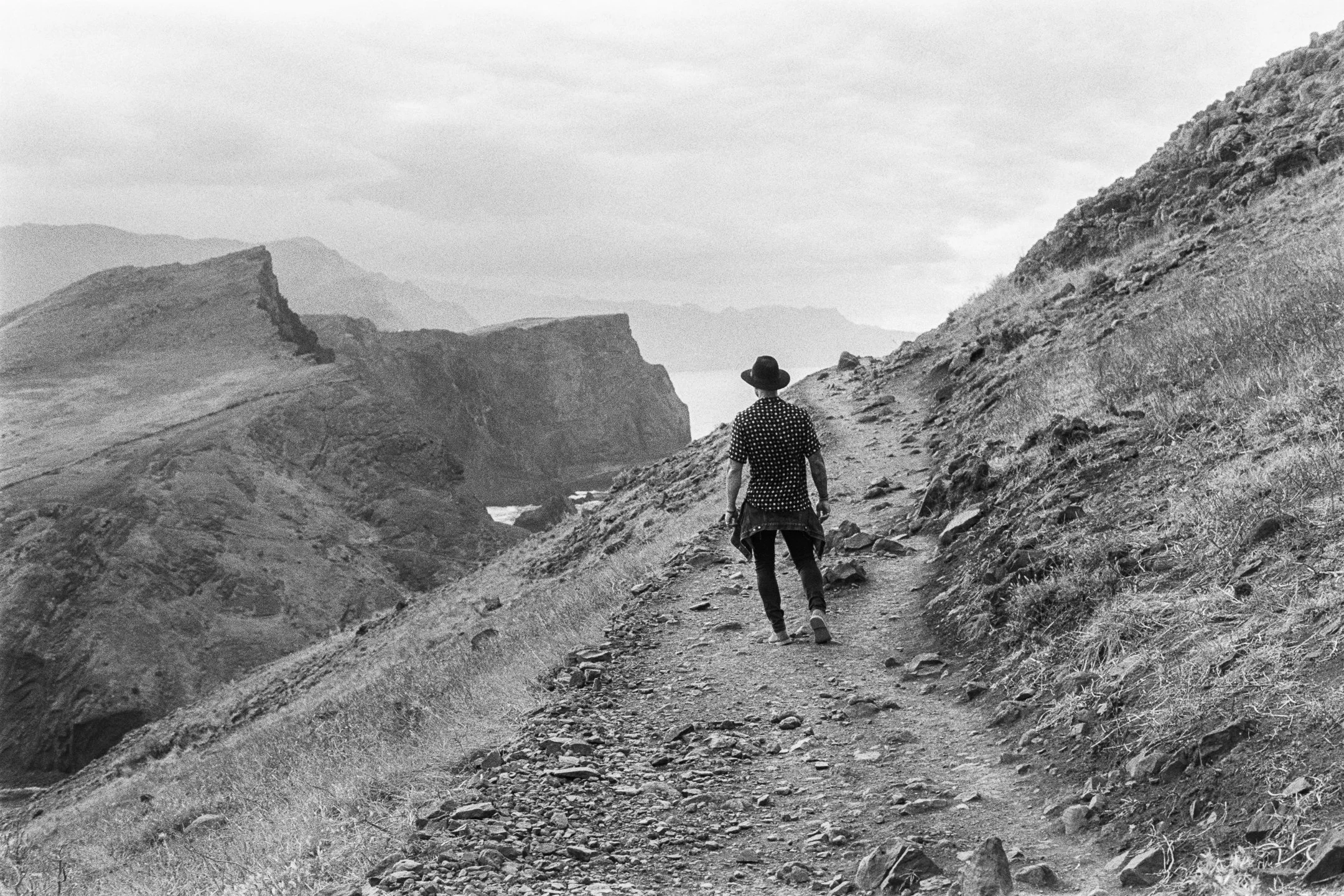 A person walking along a mountain trail through a rugged landscape with mountains in the distance, black and white photograph. photo by Bruno Novais