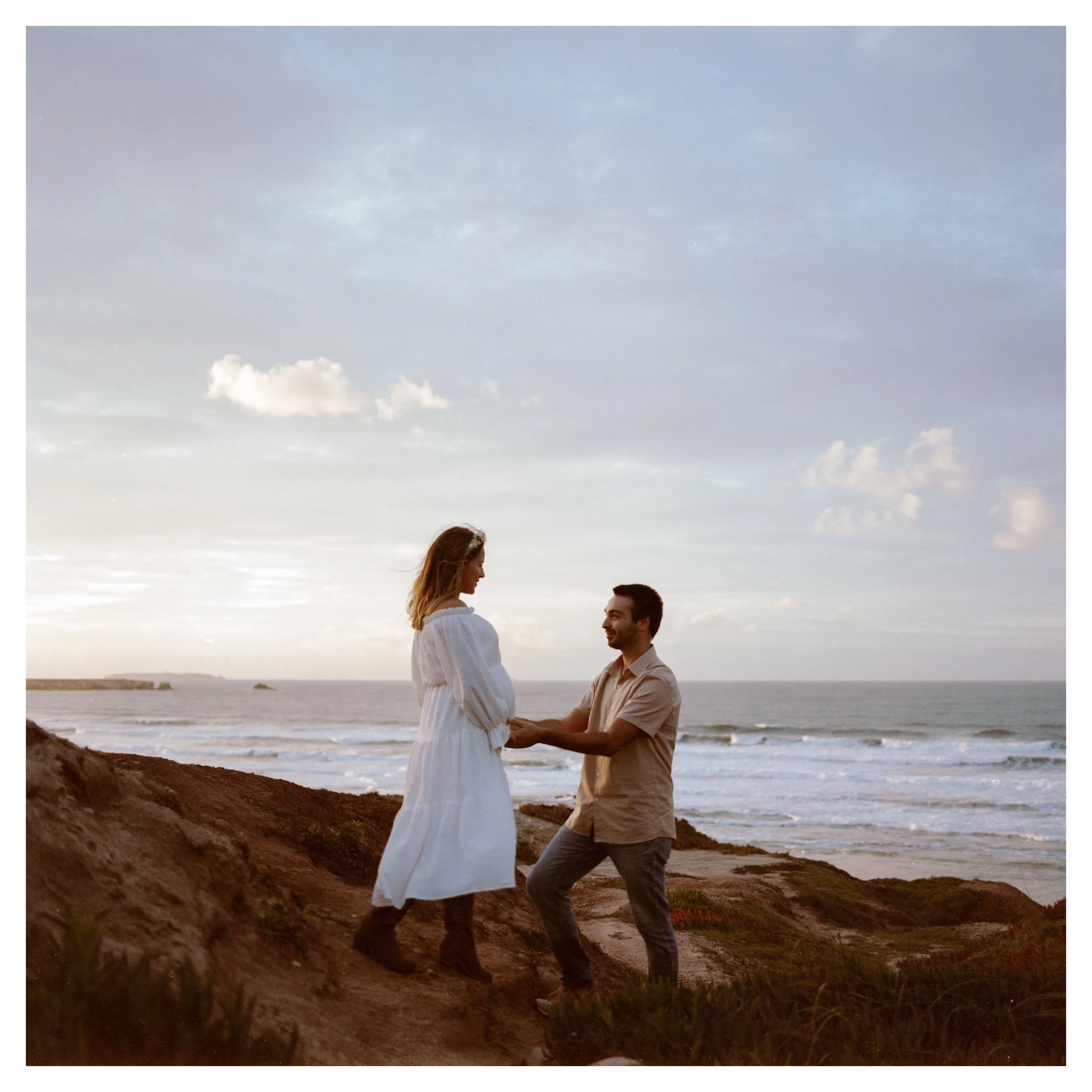 A couple with the man proposing a marriage proposal on a beach at sunset. The woman is wearing a white dress, and the man is in a beige shirt and gray pants. They are standing on a rocky area with the ocean and cloudy sky in the background.