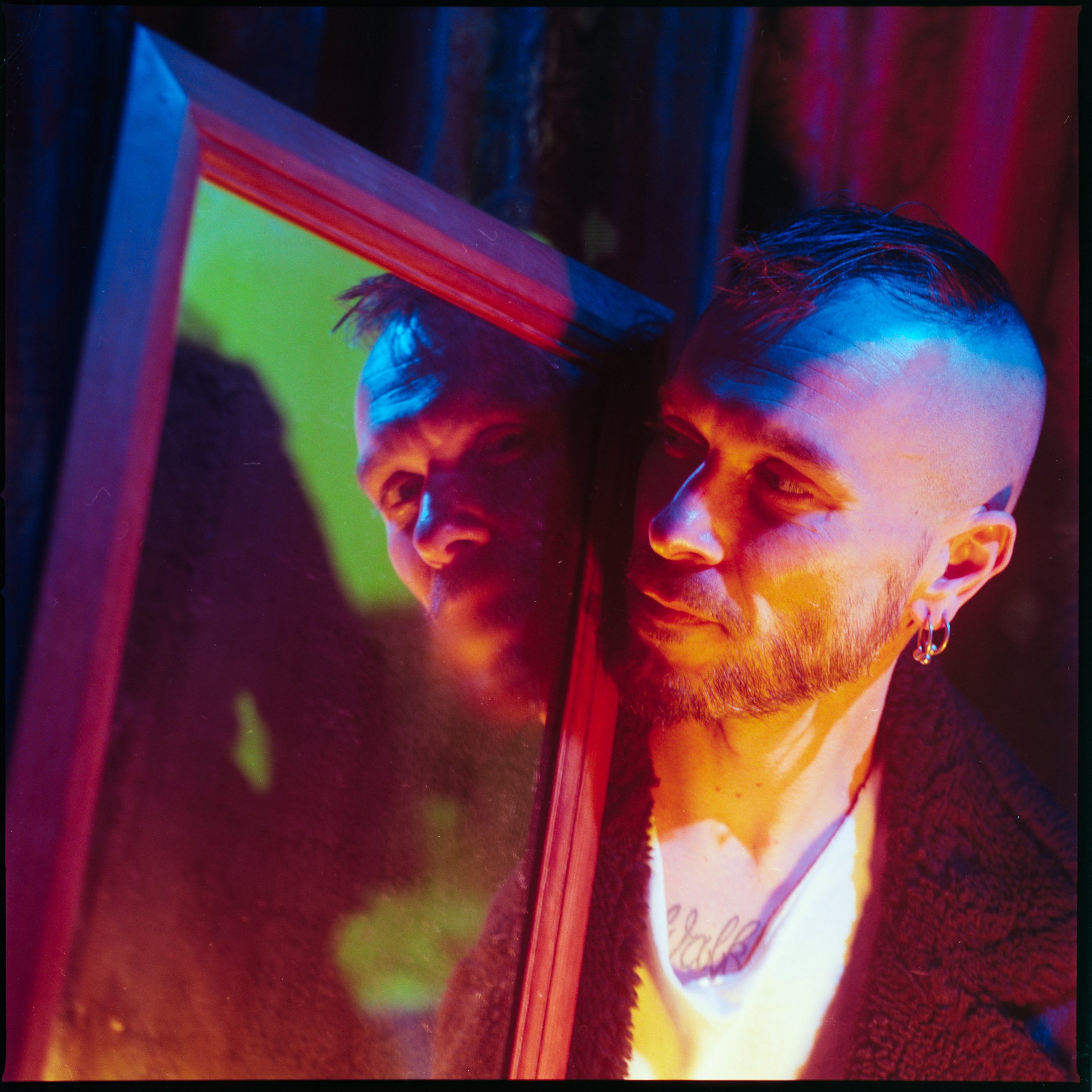 A person with short hair and earrings looks into a mirror, with colorful lighting reflecting on their face and reflection.